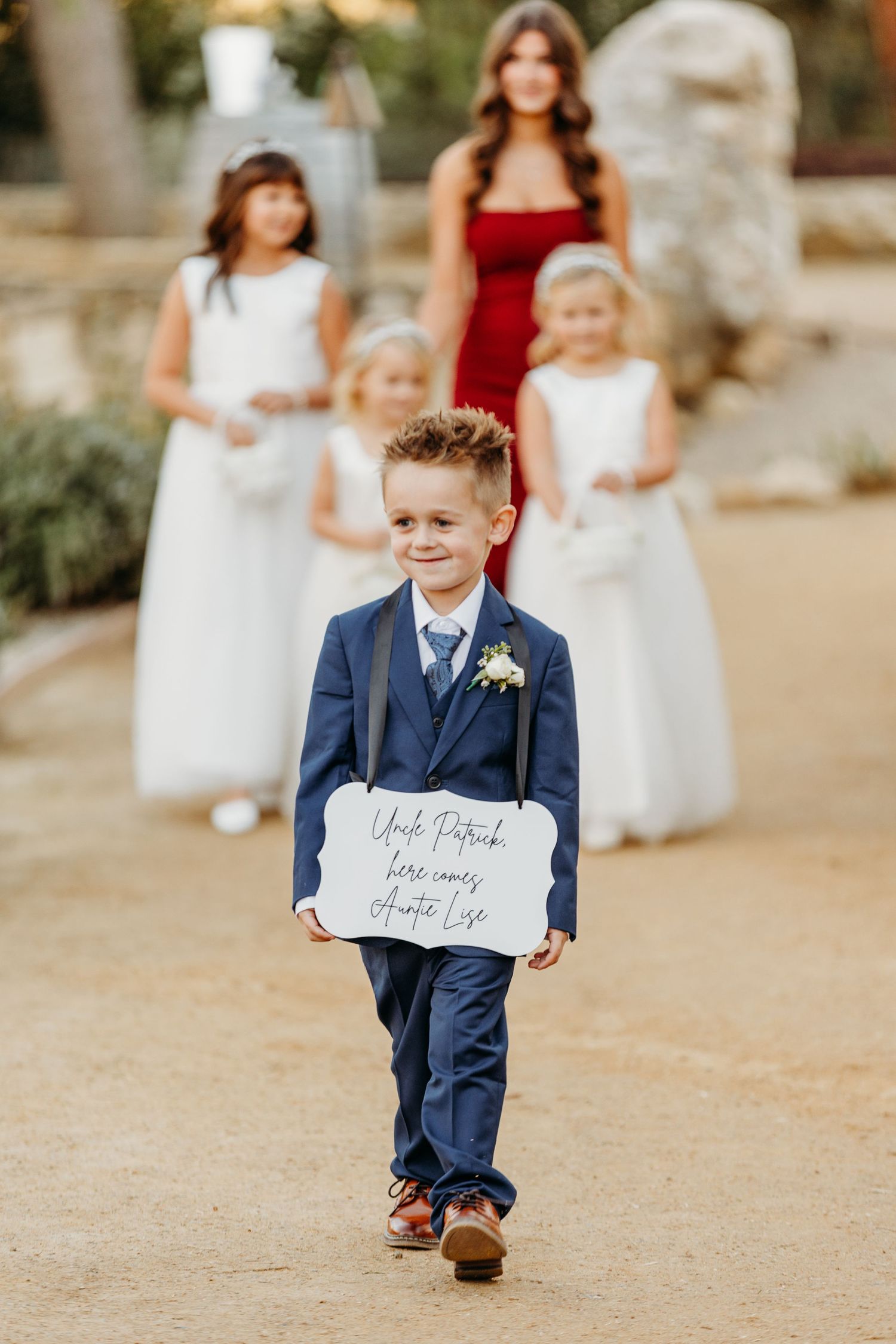 Young ring bearer in navy suit walks down aisle carrying sign at outdoor wedding ceremony.