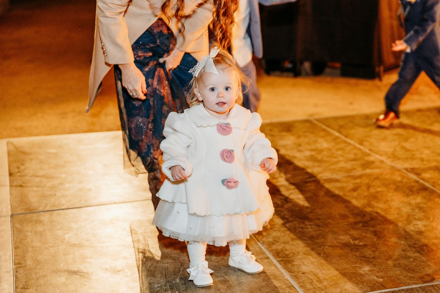 Small child in white dress dancing on wooden floor at reception.