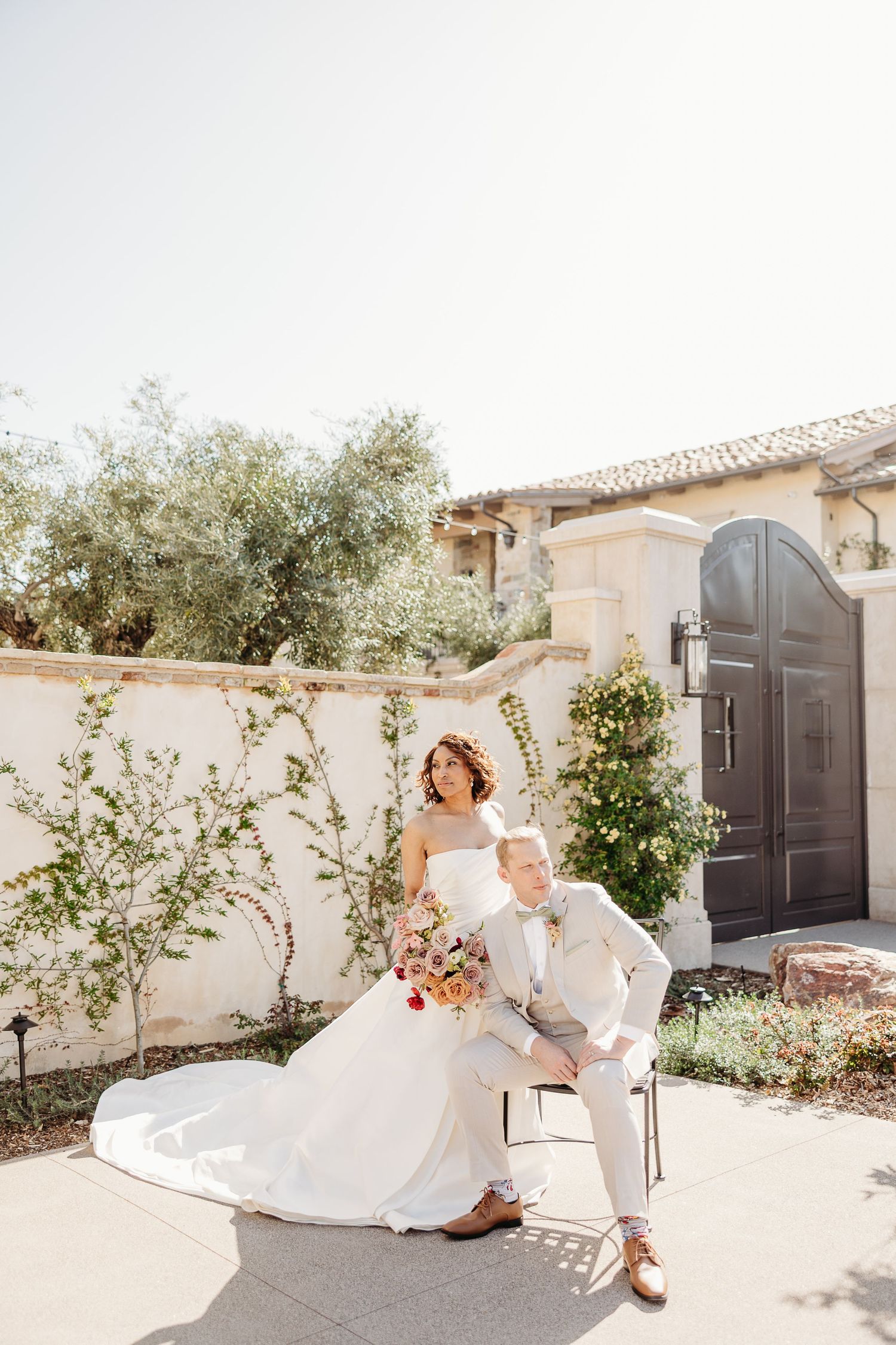 Couple in elegant wedding attire share intimate moments in a sun-drenched Mediterranean courtyard with white walls.