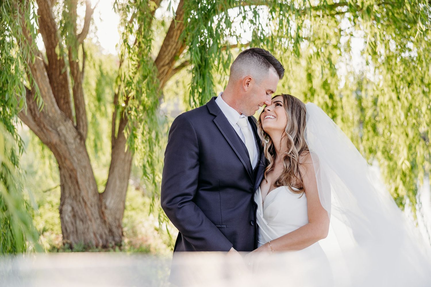 Wedding couple embraces under weeping willow trees in romantic garden setting.