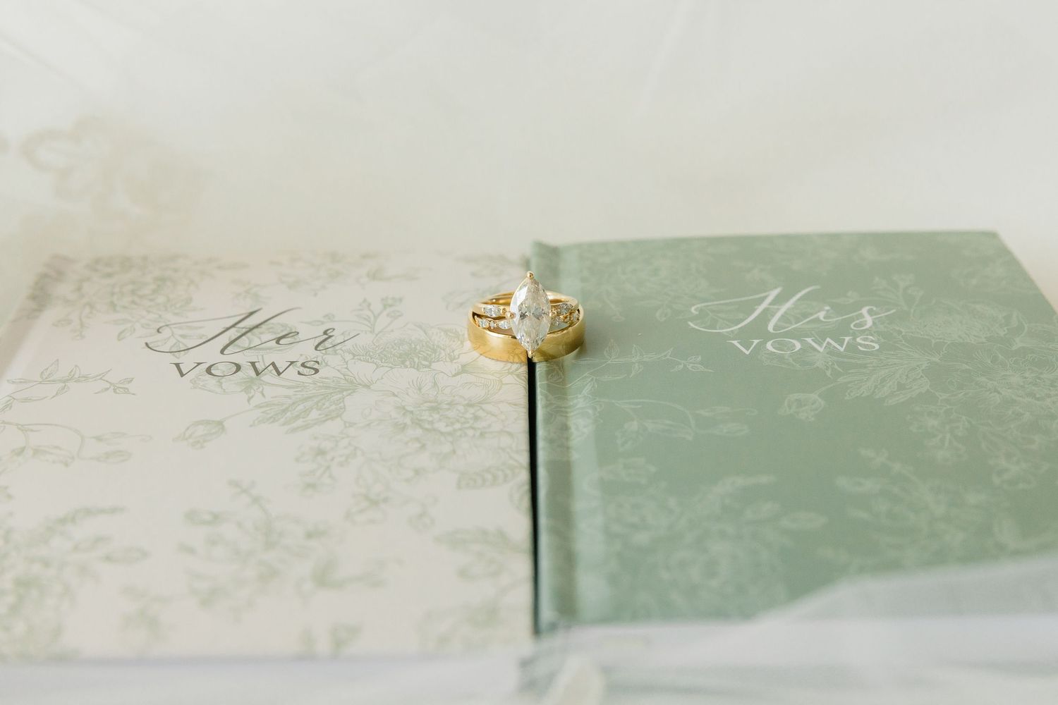 Gold engagement ring resting on Her Vows and His Vows green floral wedding vow books.
