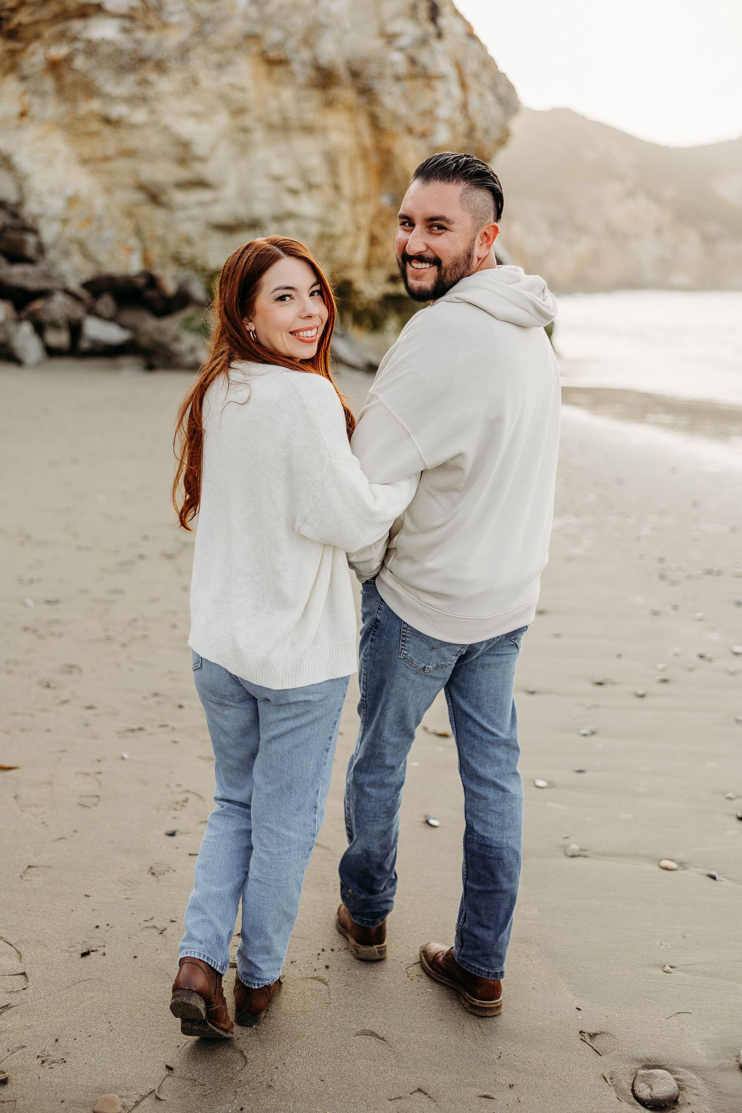A couple wearing matching white sweaters and jeans walk together on a beach with cliffs in the background.