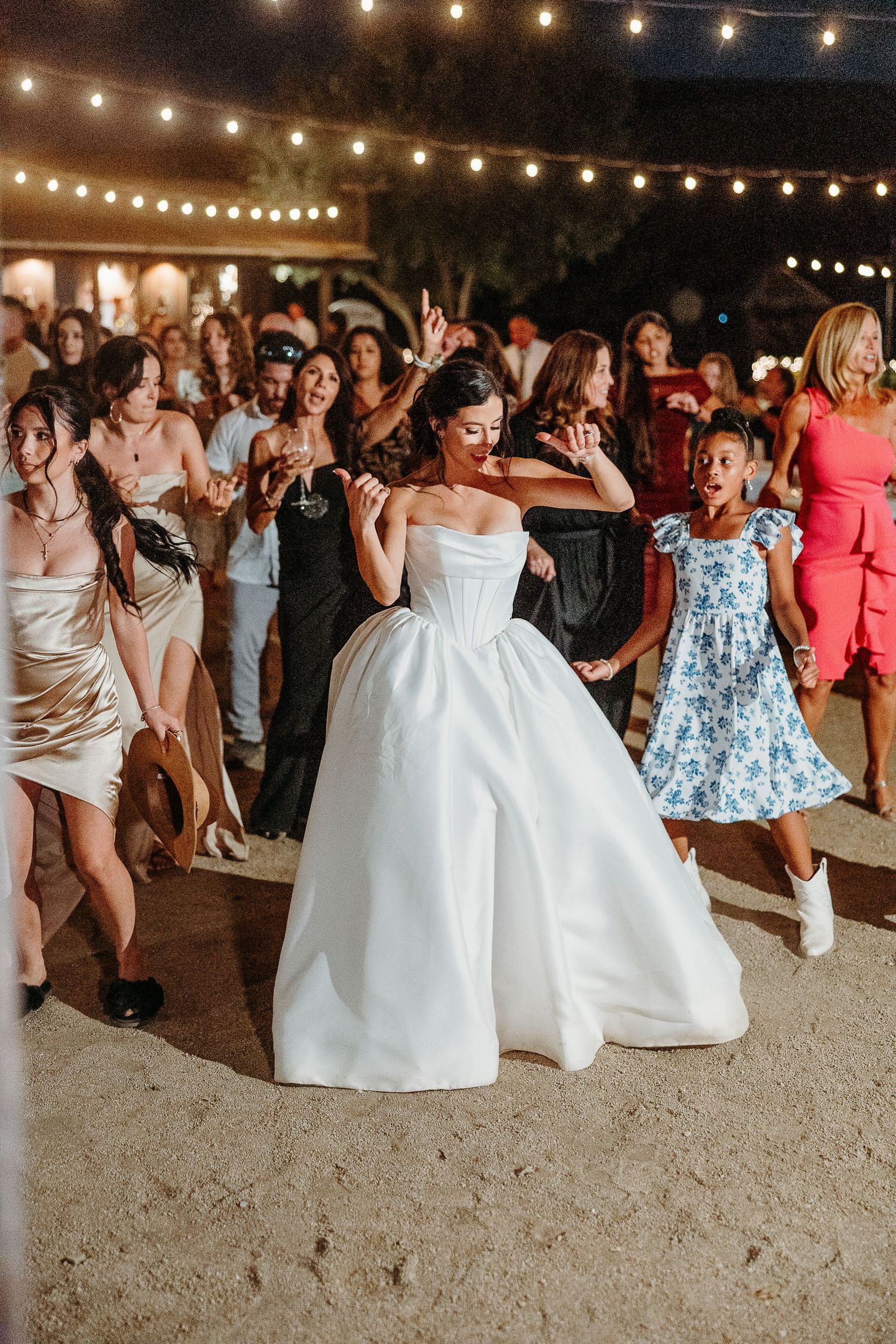 Wedding reception dance floor filled with guests in white dresses under string lights.