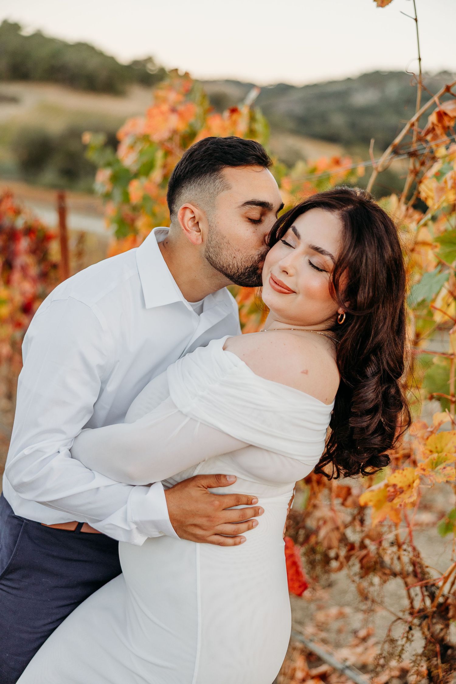 A romantic couple embraces among autumn vineyard vines with warm orange and golden colors at sunset.