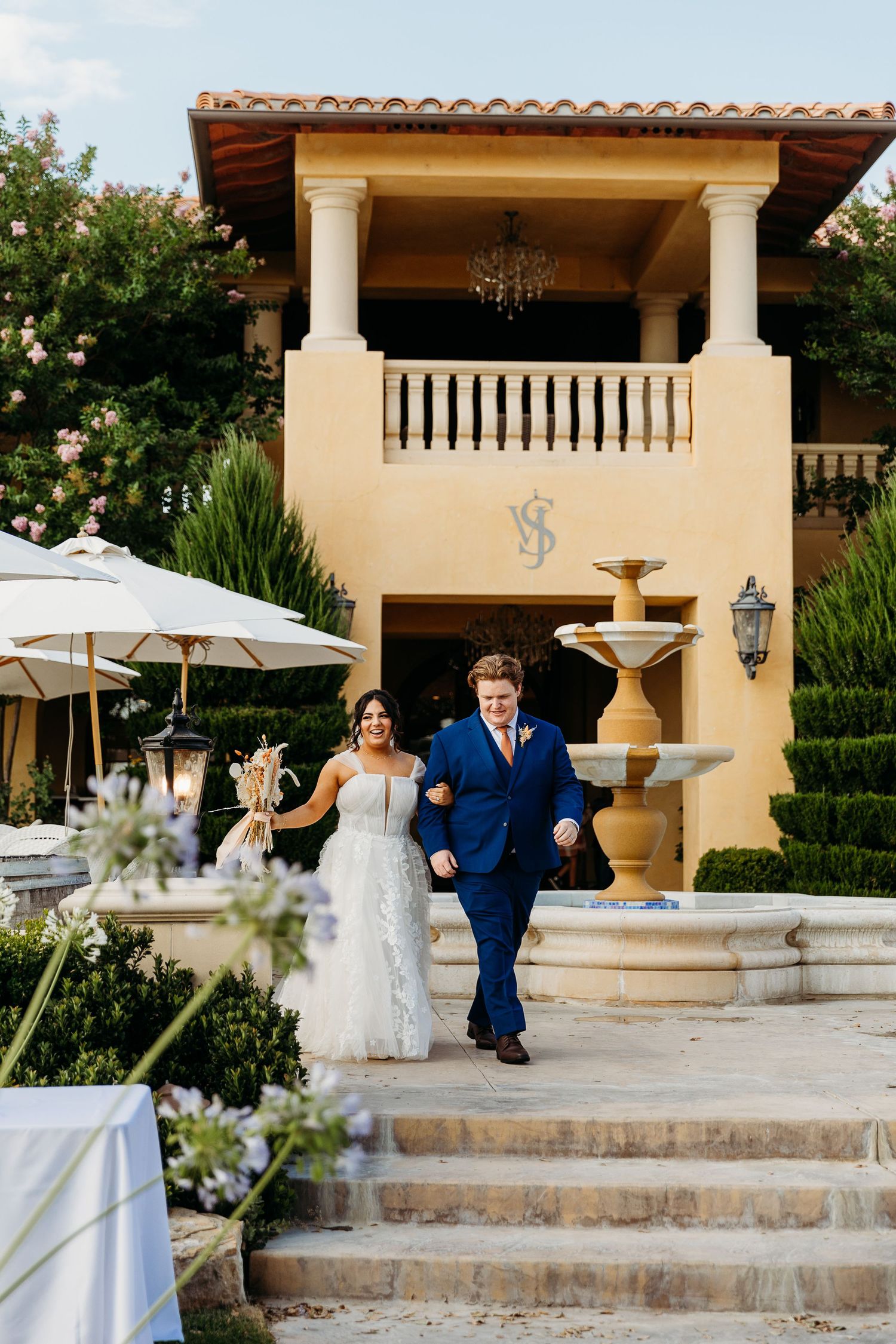 Elegant couple descending stone steps at Mediterranean villa with fountain and white umbrellas.