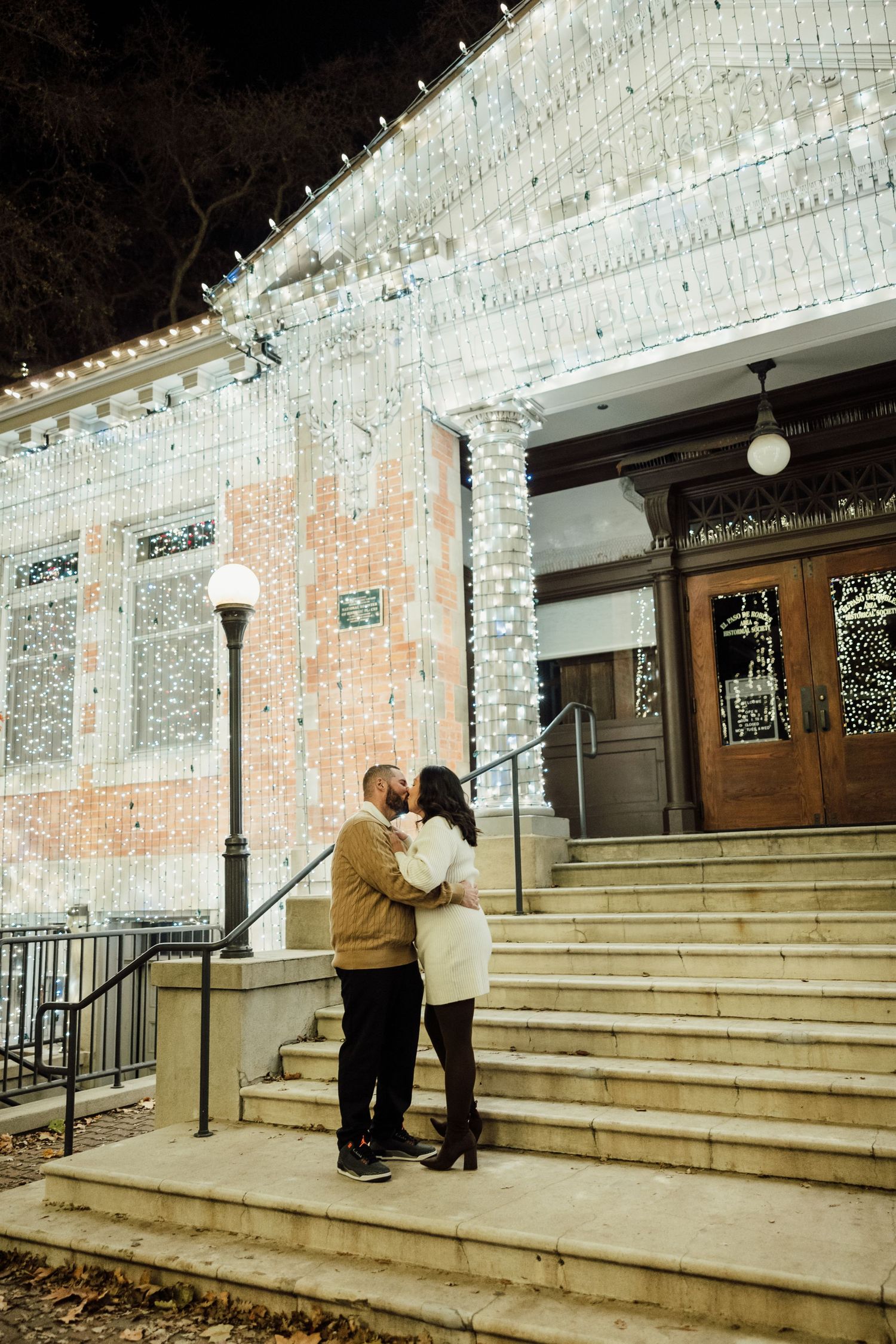 Two people stand on concrete steps outside a building entrance with white lights decorating the exterior.