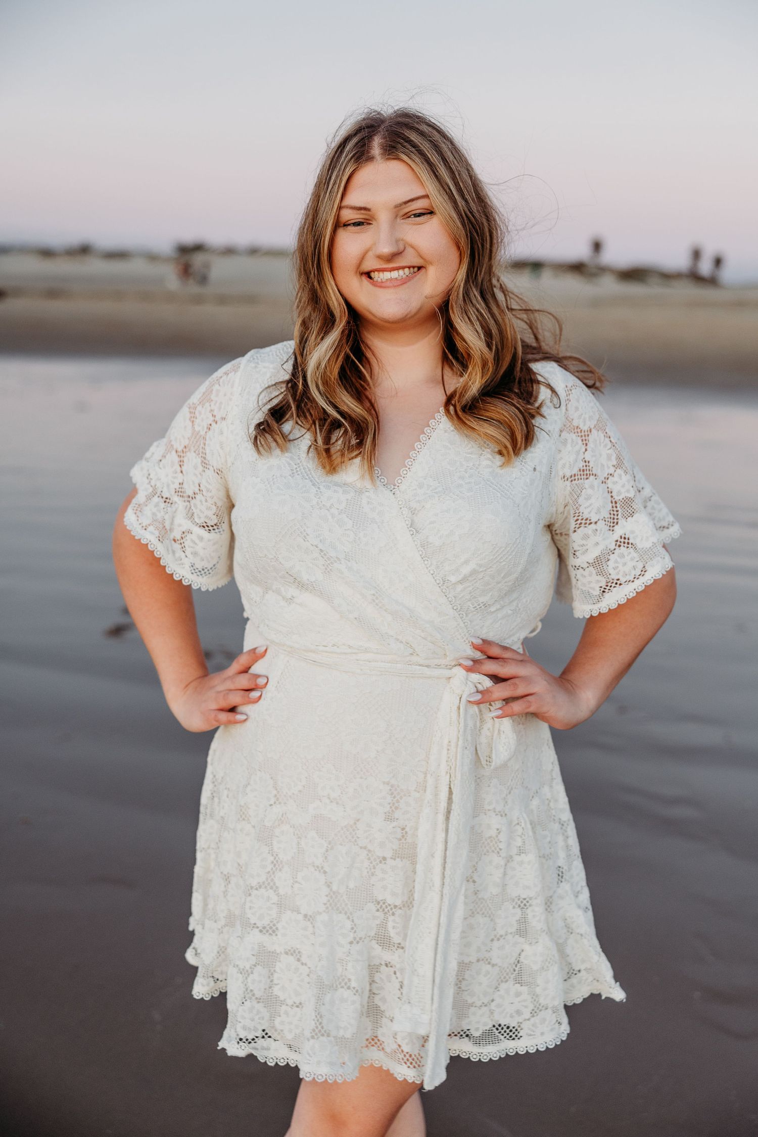Someone wearing a white lace wrap dress poses confidently on a beach at sunset.