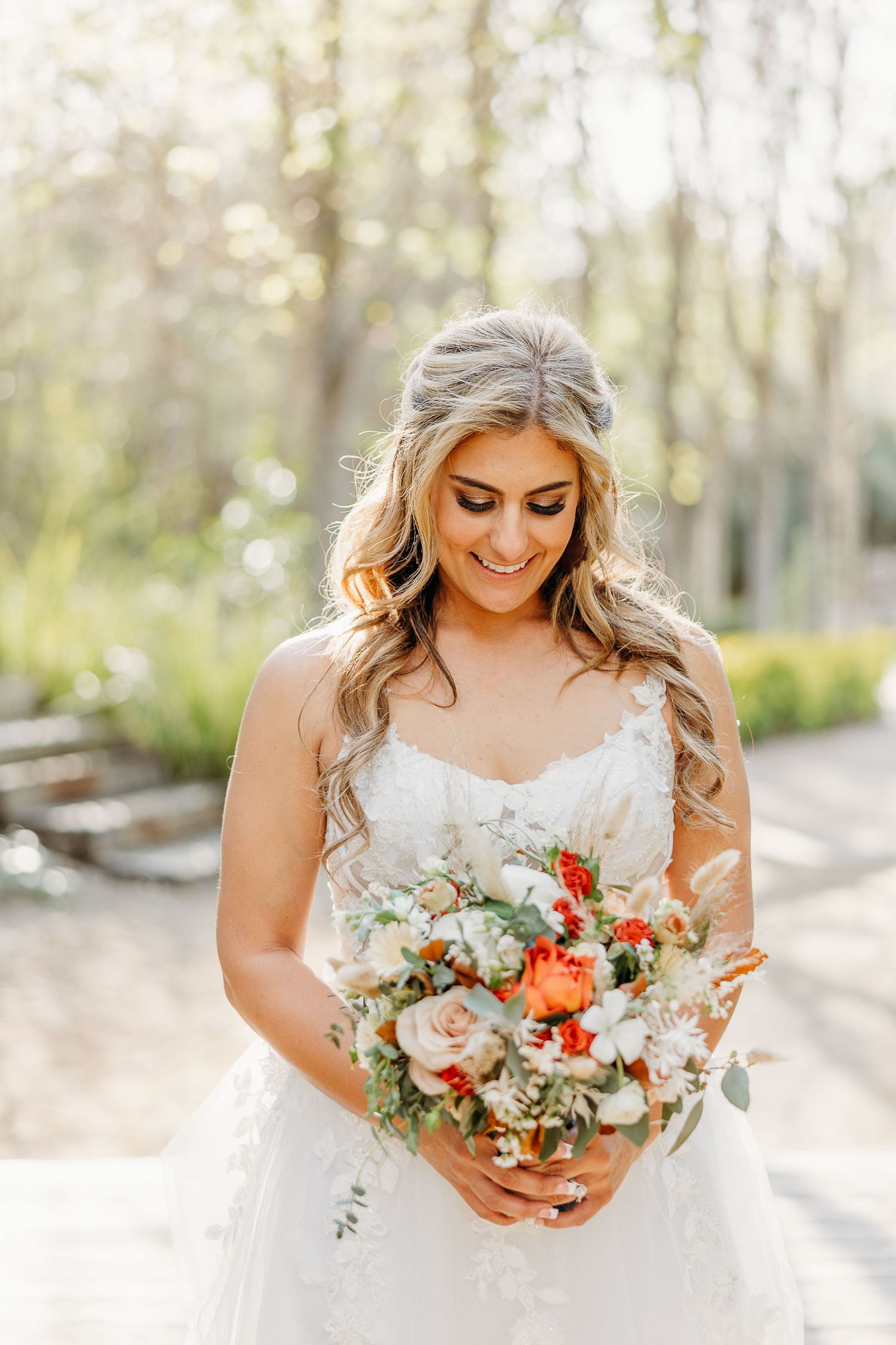 Bride in white strapless wedding dress holds beautiful bouquet with peach and coral flowers on sunny outdoor path.