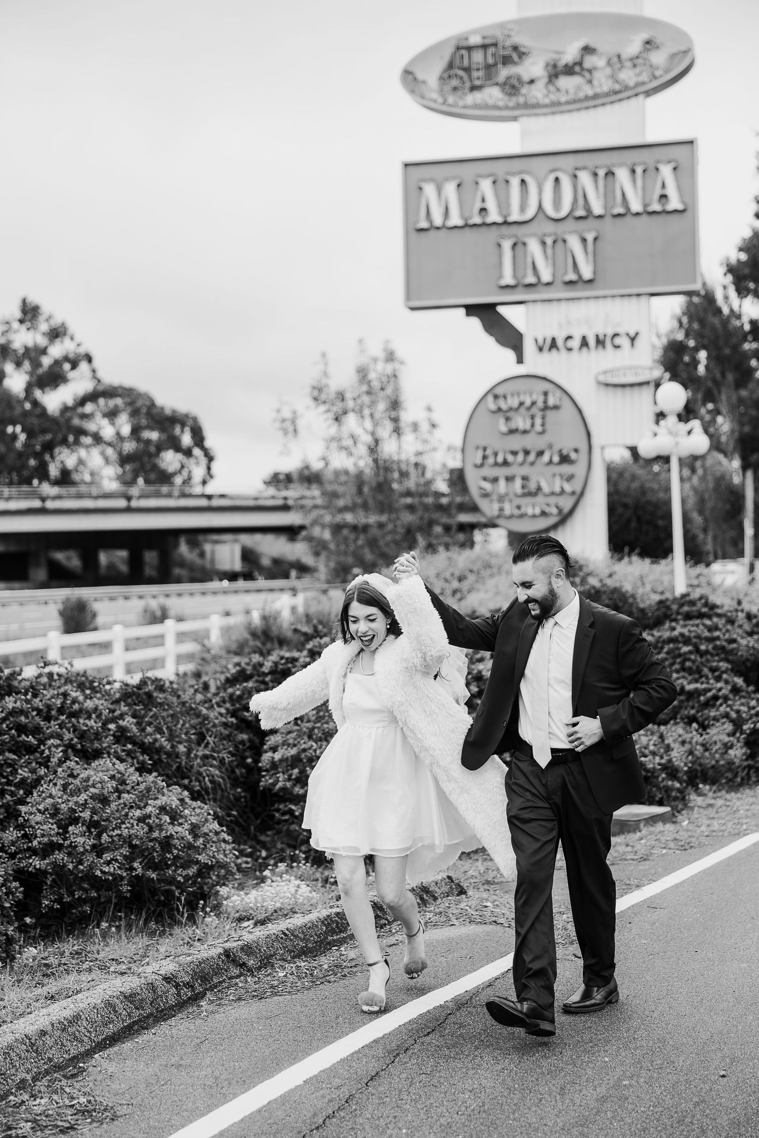 A joyful couple runs along a road in front of the Madonna Inn sign in a vintage black and white photograph.