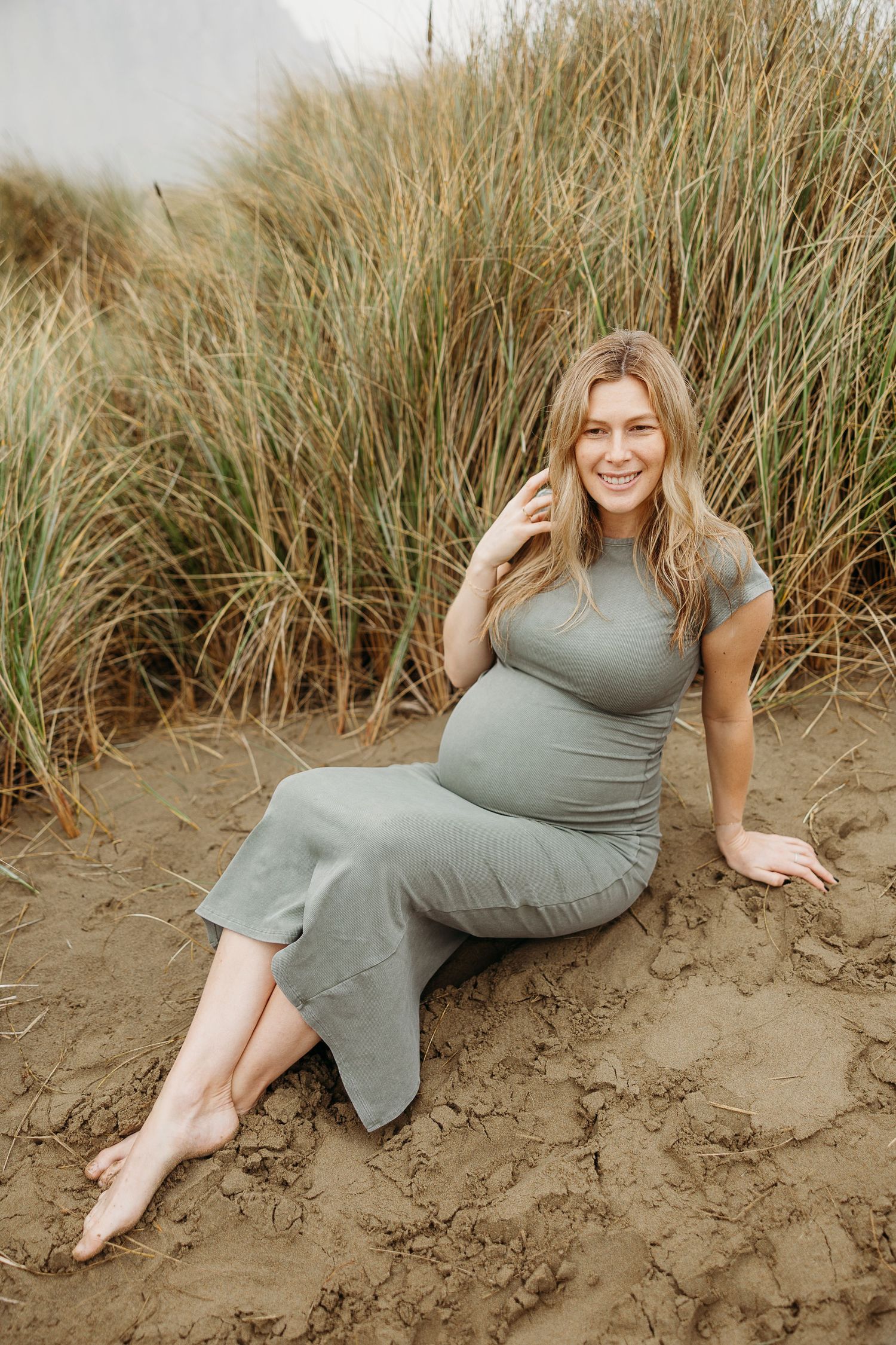 Casual portrait of expectant mother in gray dress sitting on beach with natural grass background.