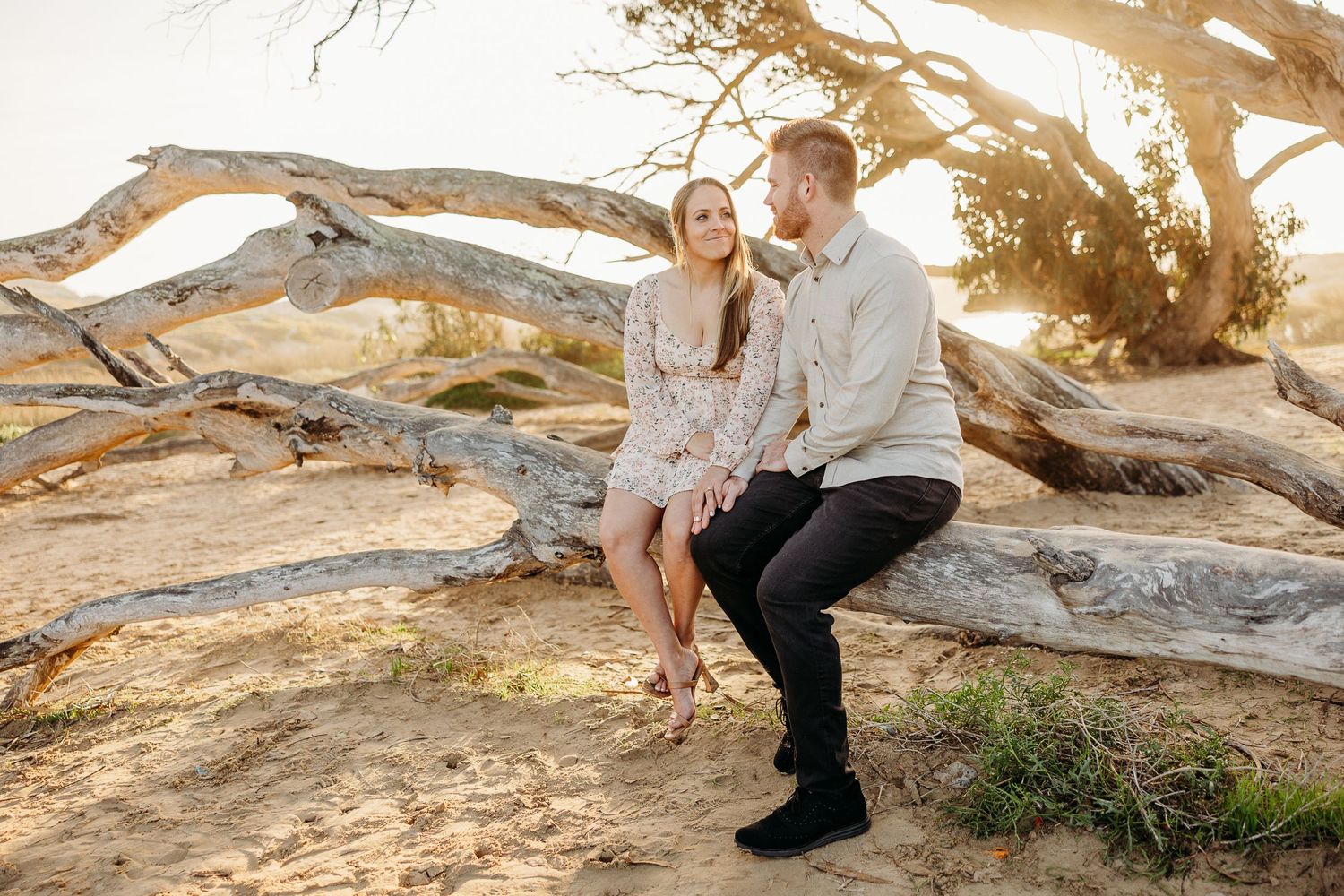A couple sits together on driftwood at sunset in a coastal setting.