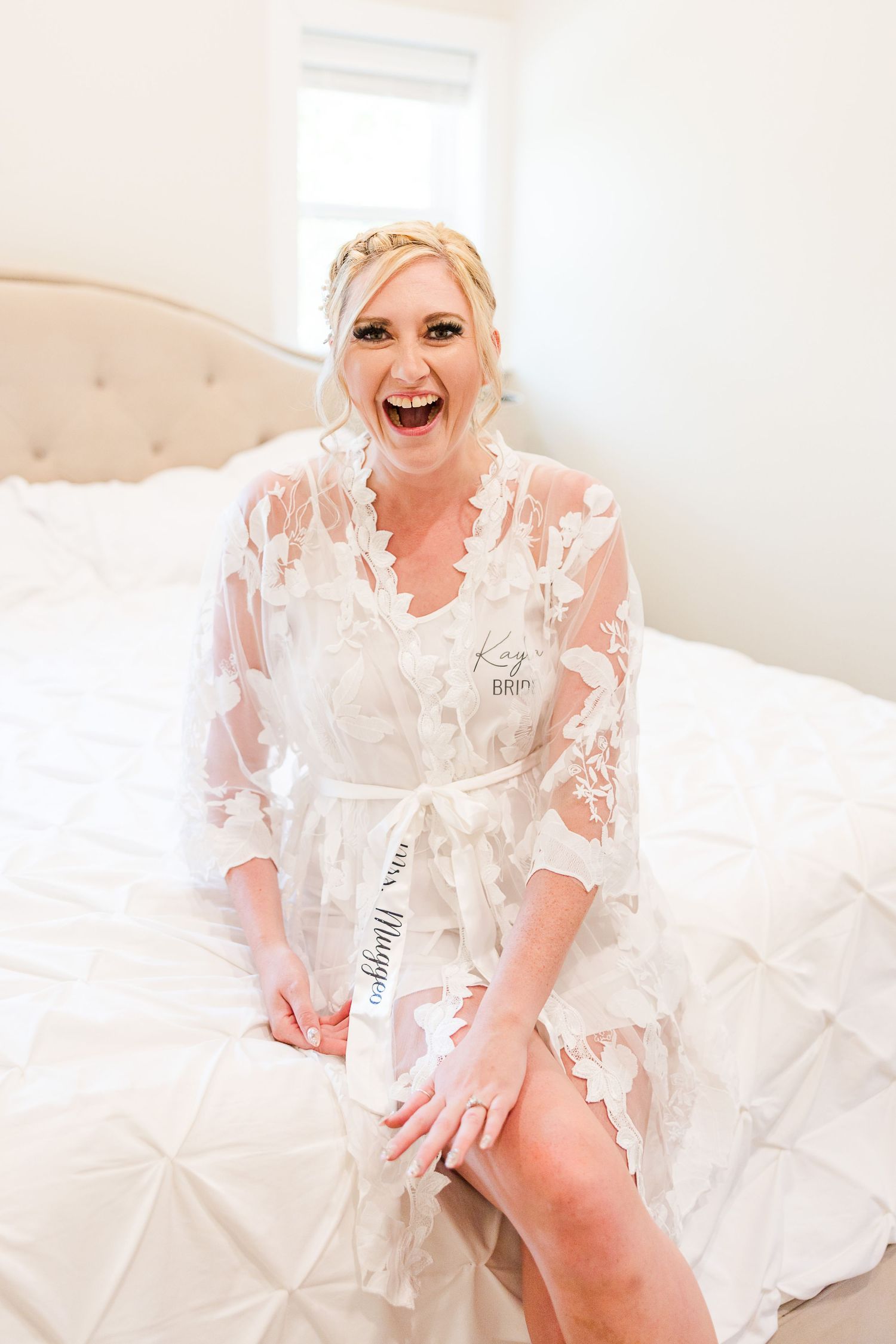 Person wearing white lace robe sits on bed laughing during wedding preparations.