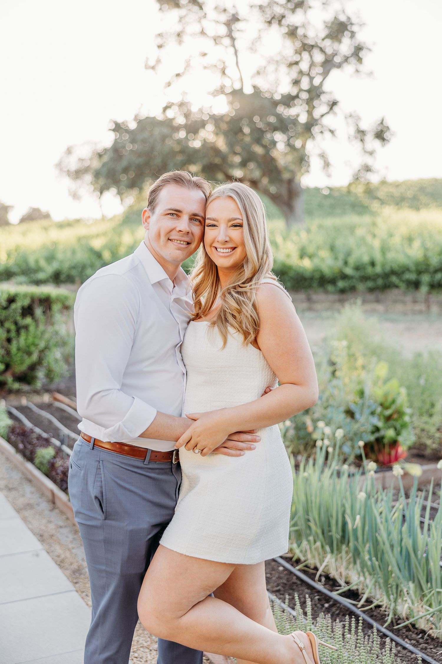 Couple embraces playfully in a garden setting with raised plant beds during golden hour sunset.