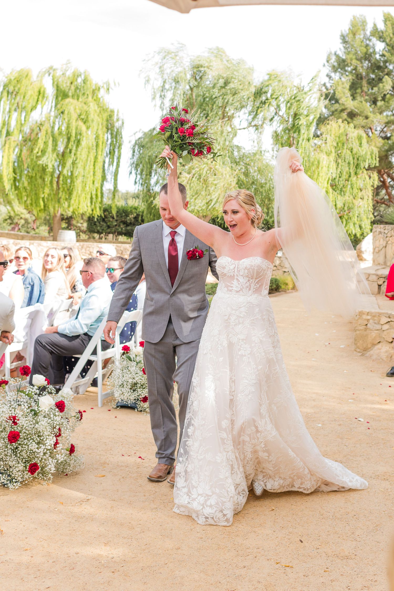 Newlyweds celebrating with raised bouquet while walking down outdoor aisle.