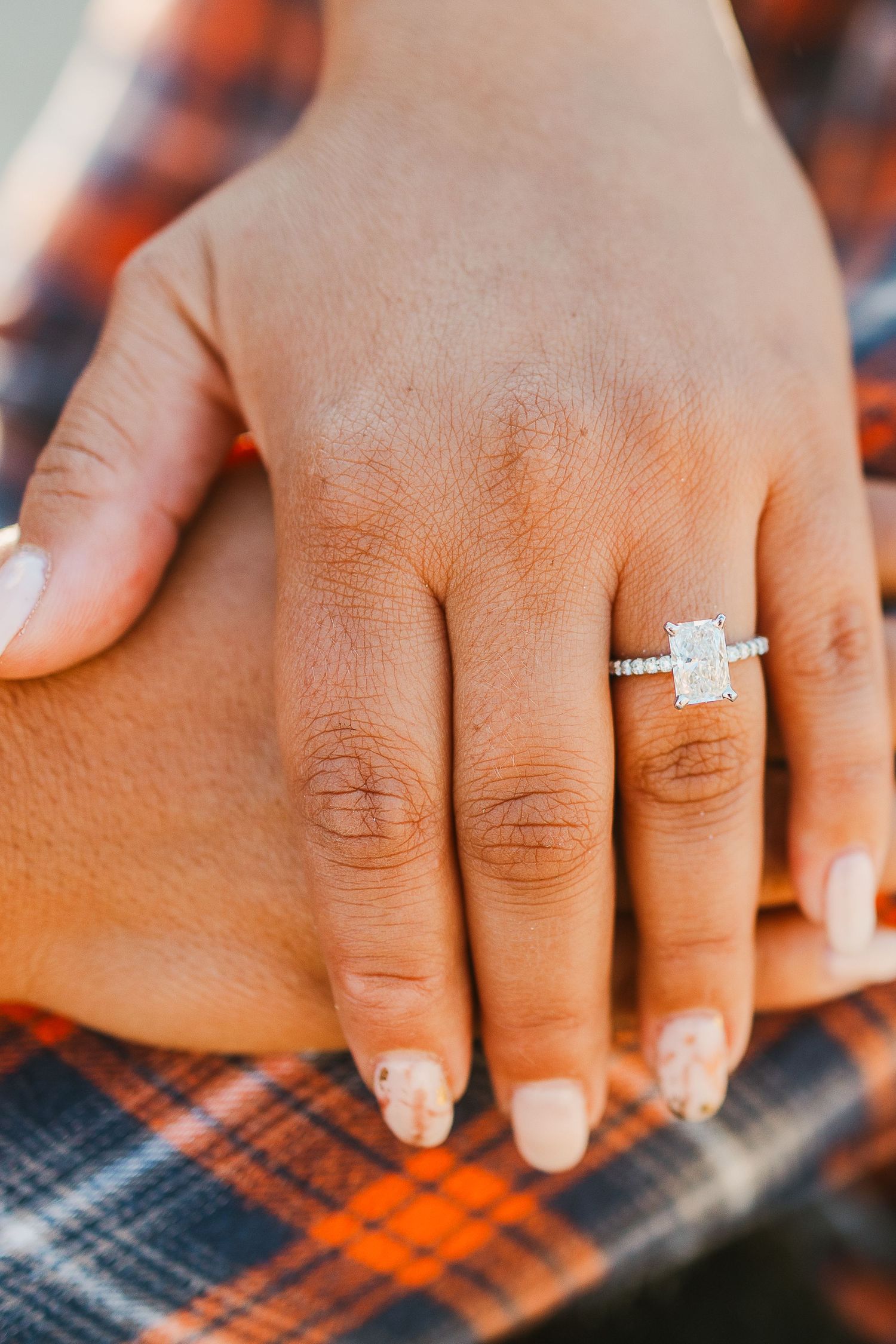 Close-up of hand wearing elegant diamond engagement ring against plaid fabric background.