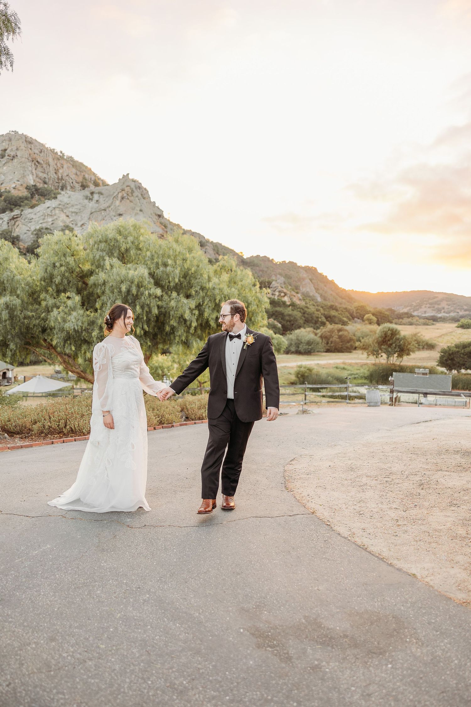 Couple in wedding attire walking along a paved path with mountains and sunset in the background.