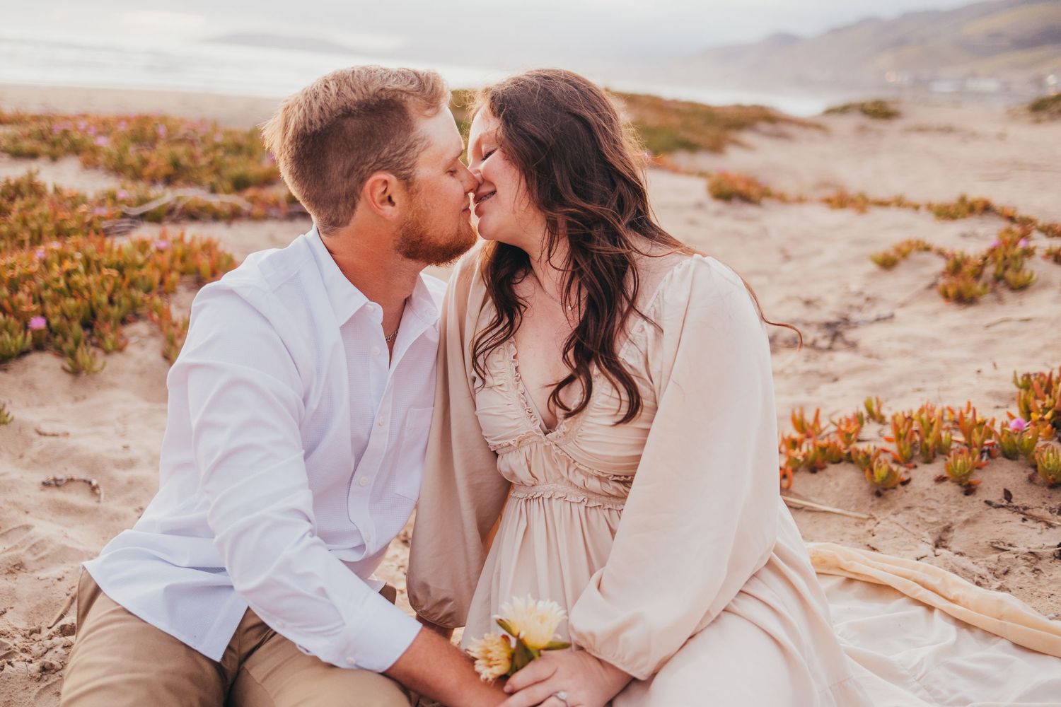 A romantic couple shares an intimate moment at sunset on a coastal hillside with desert flora.