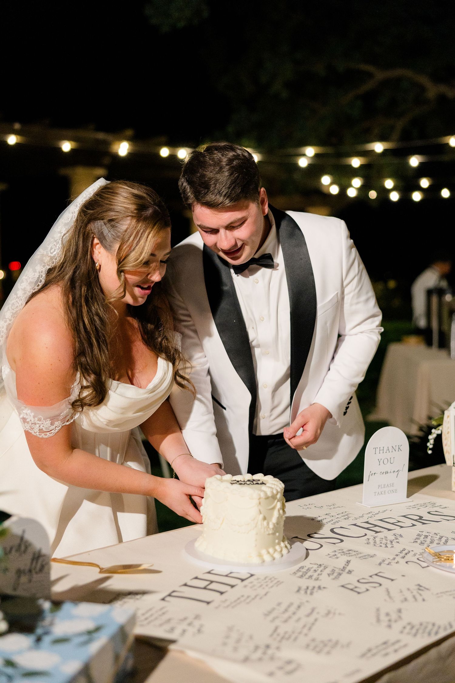 Bride and groom laugh while cutting white wedding cake at outdoor evening reception.