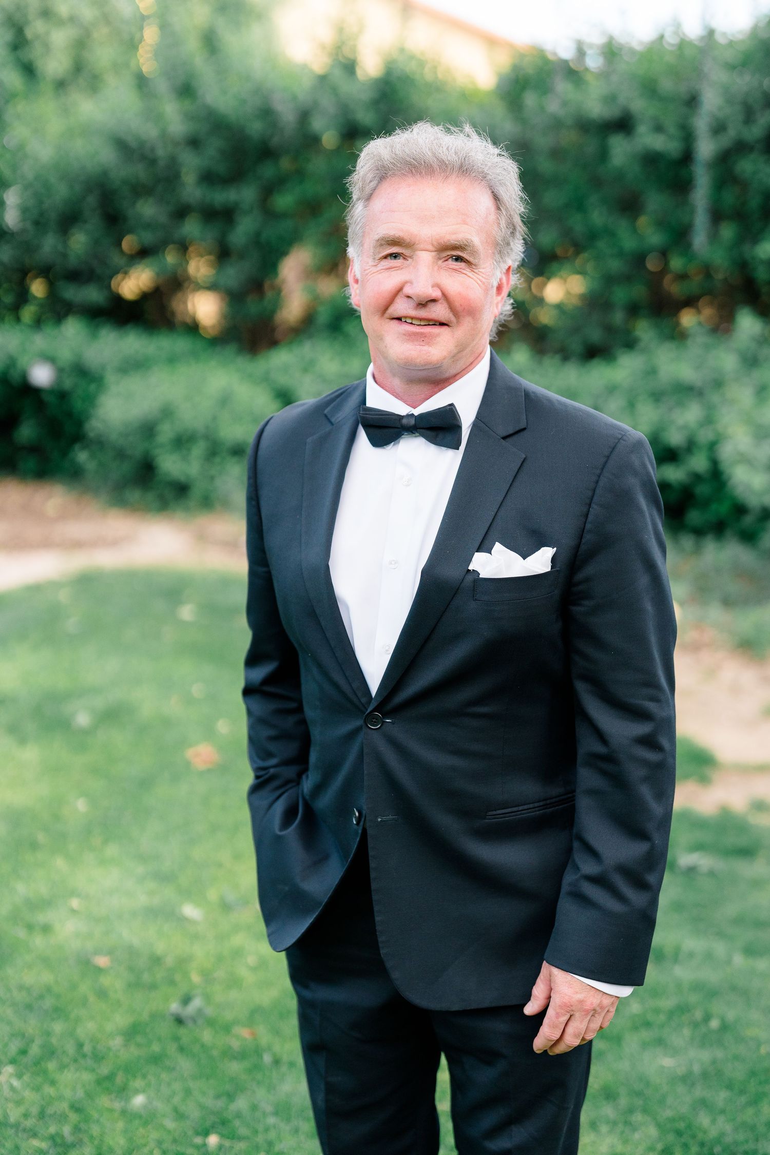 Elegant older man in black tuxedo with bow tie smiling outdoors.
