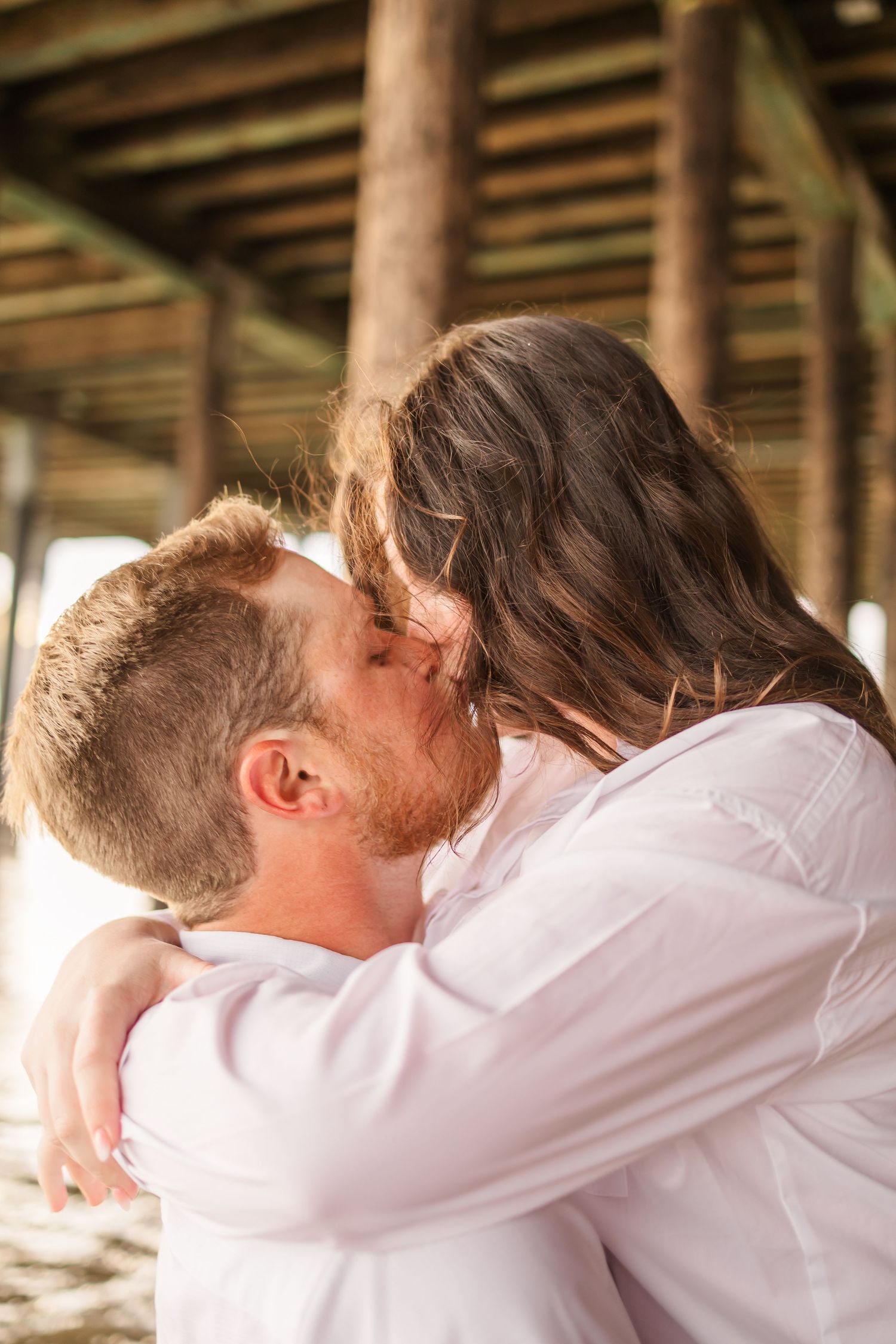 Romantic embrace captured under pier during beach photoshoot.
