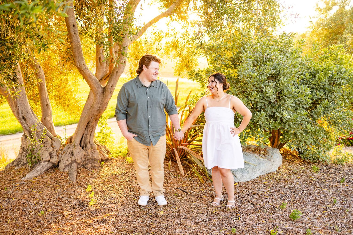 Happy couple walking together on gravel path surrounded by trees in warm evening light.