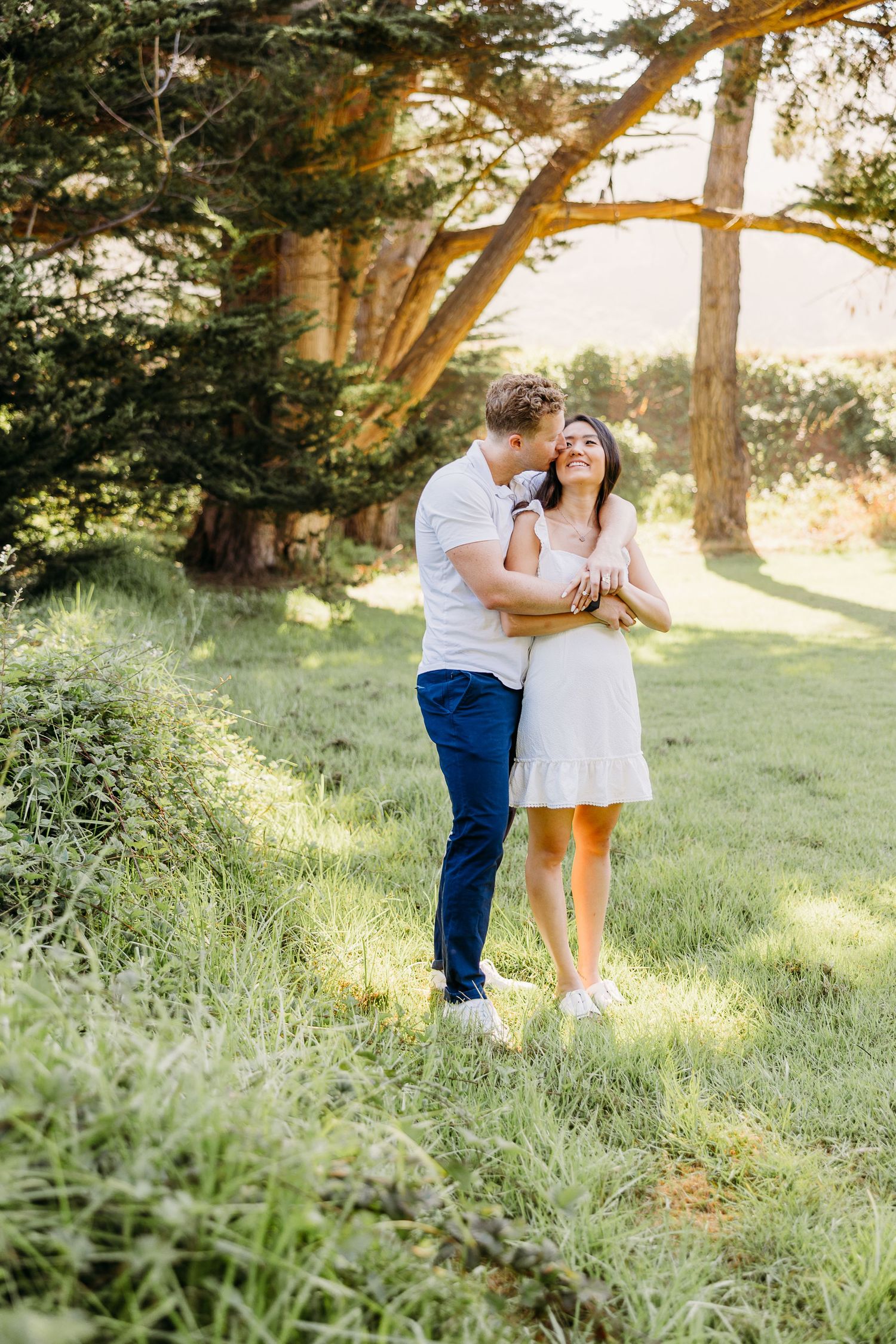 A couple shares an intimate moment on a grassy hillside with tall trees in the golden afternoon light.