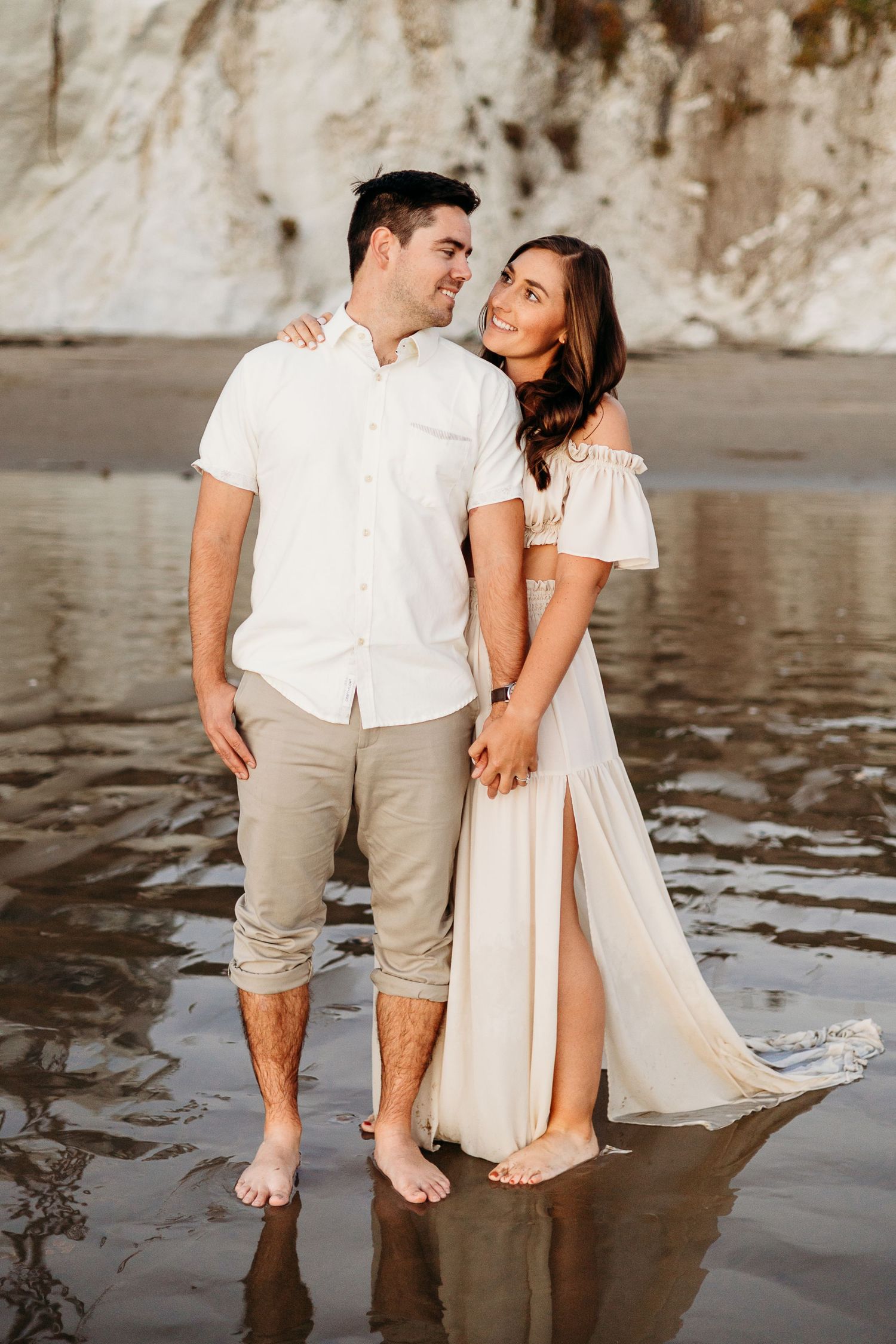 A couple in coordinated outfits standing close together on a reflective beach with white cliffs behind them.