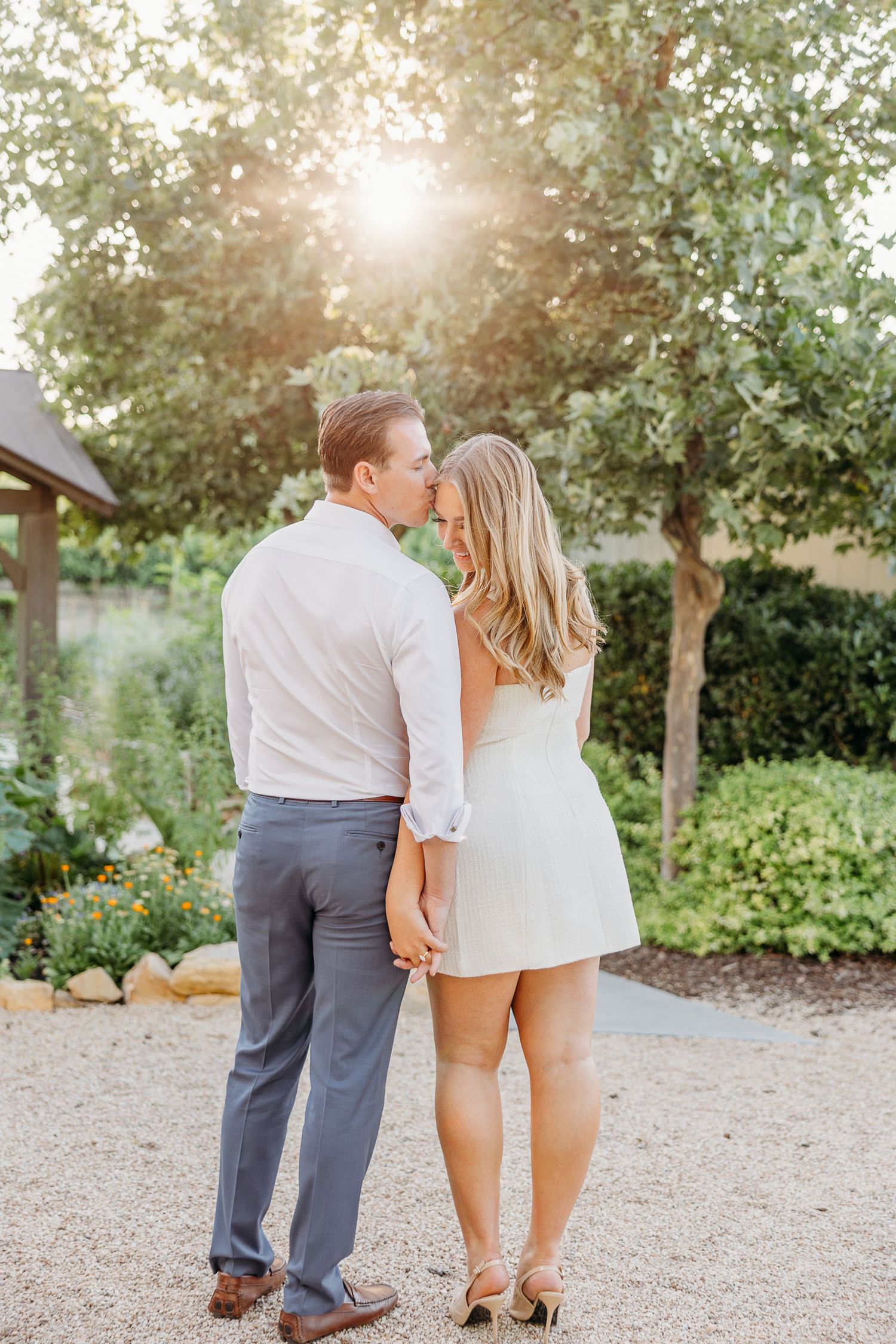 A couple walking together on a gravel path surrounded by trees and greenery at sunset.