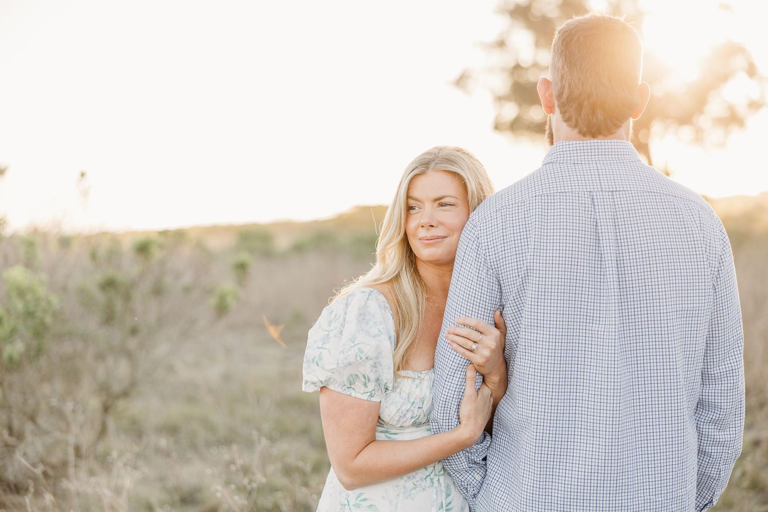 A loving couple embraces at sunset in a romantic meadow wearing blue and white outfits for their engagement photos.