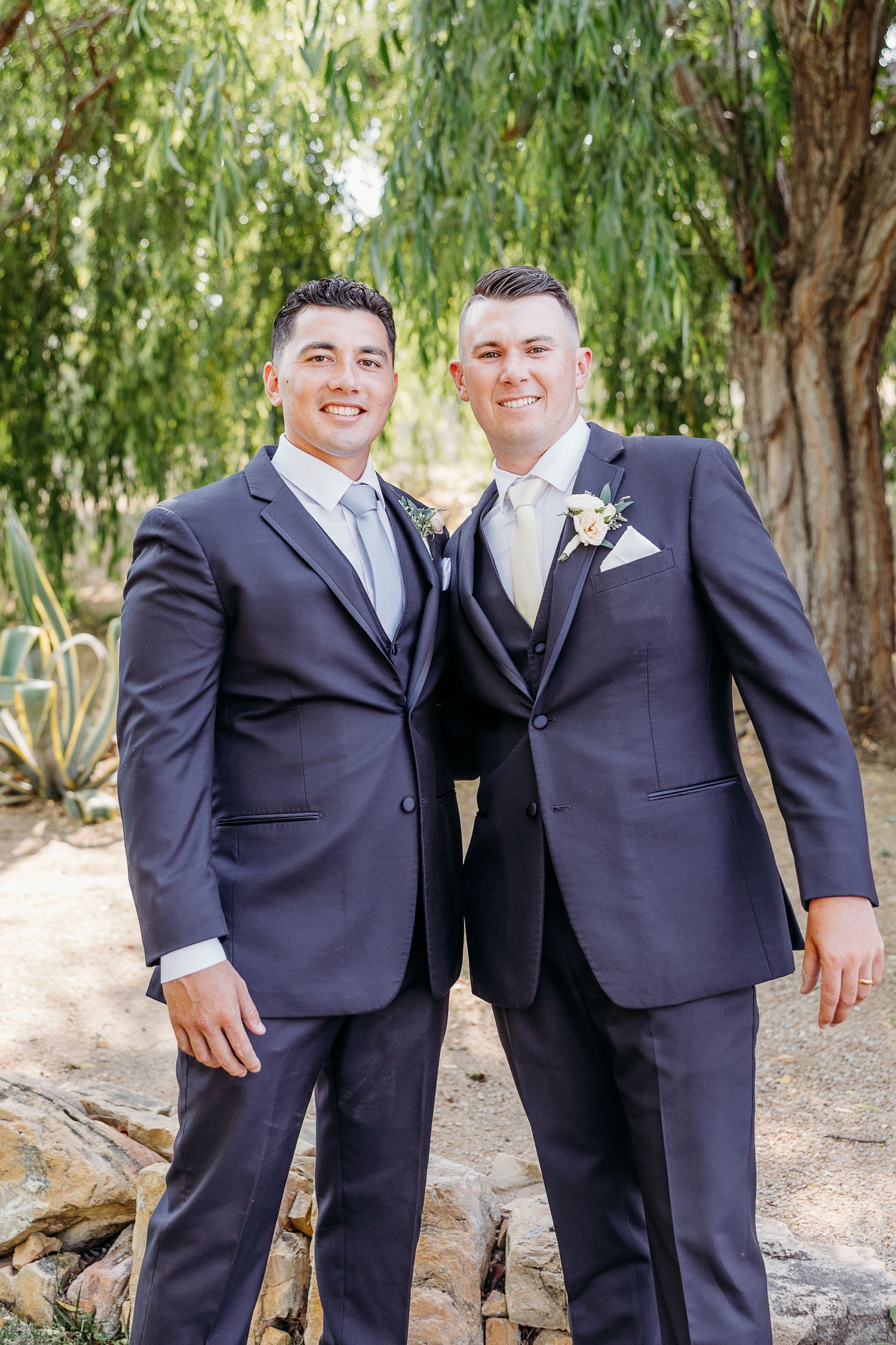 Two people wearing dark suits standing together outdoors under willow trees on a path.