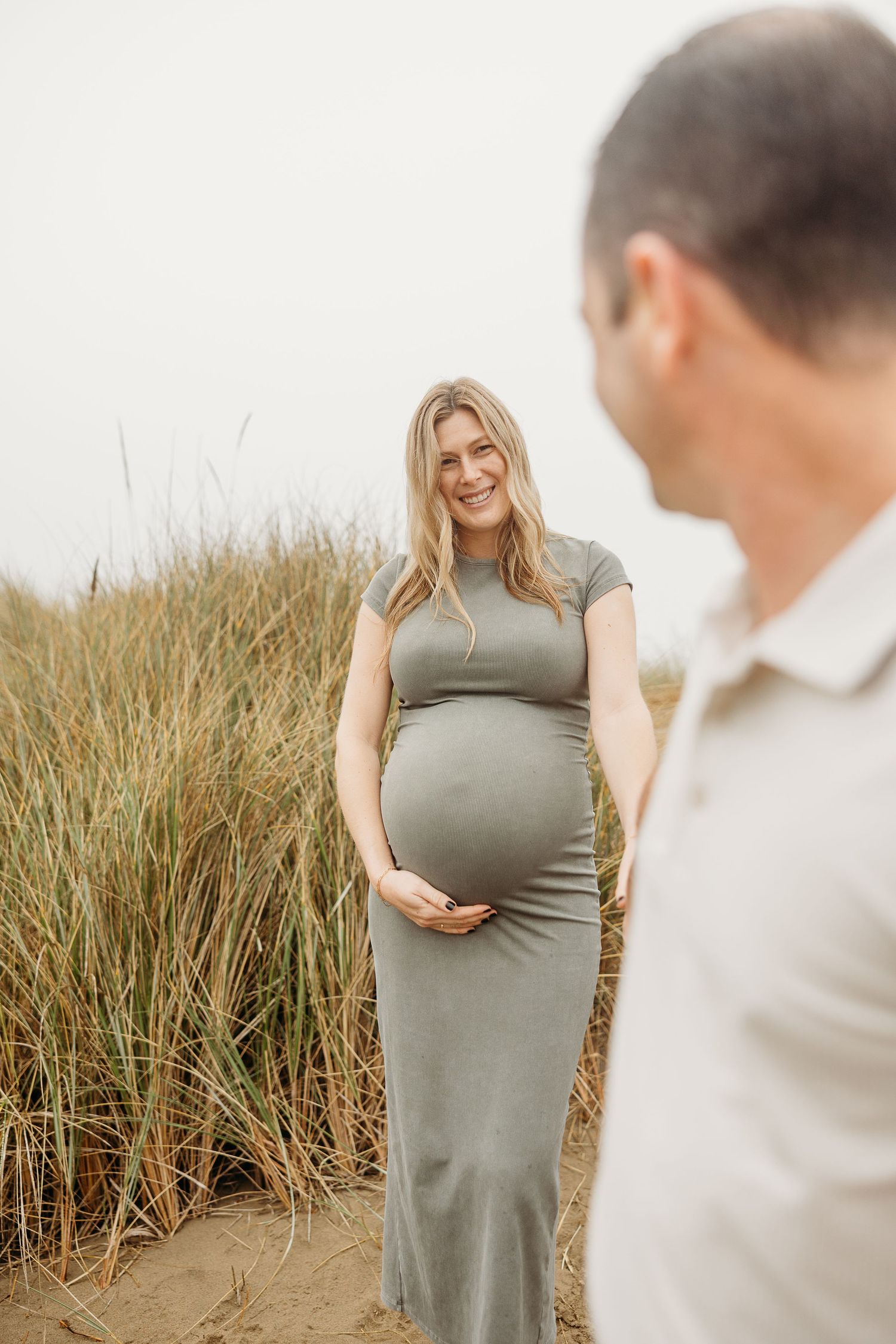 Expectant couple poses in natural setting with tall grasses as woman shows off pregnancy bump.