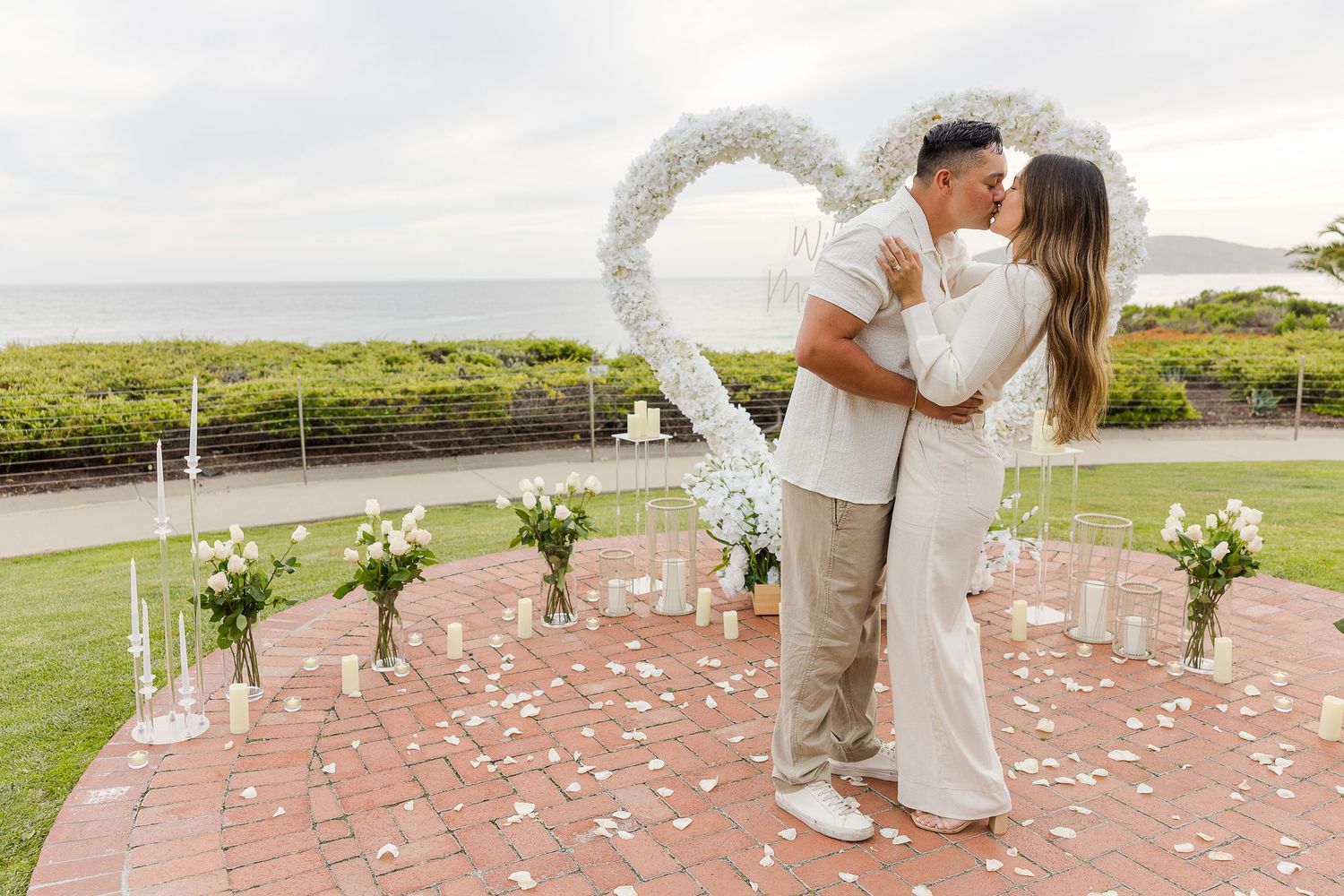 Romantic wedding ceremony scene with white floral arch and scattered petals against an ocean backdrop.