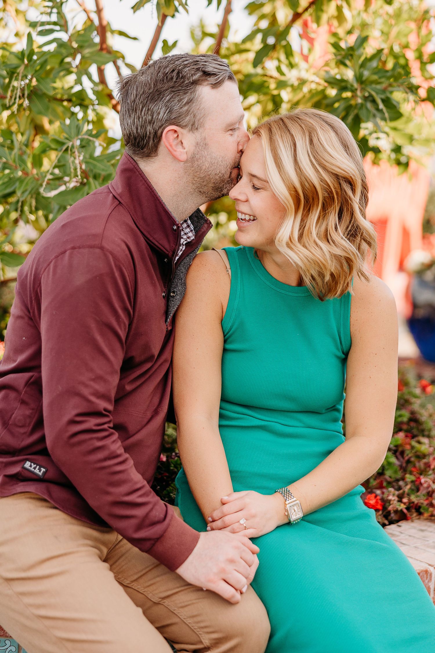 A couple sits together on a bench sharing an intimate moment in a fall garden setting with burgundy and green attire.