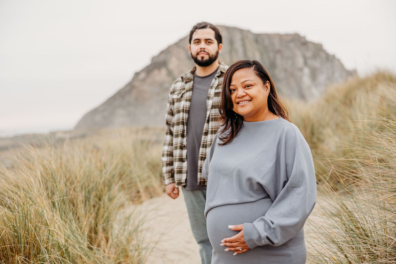 A couple stands together in tall beach grass with a mountain in the background on an overcast day.