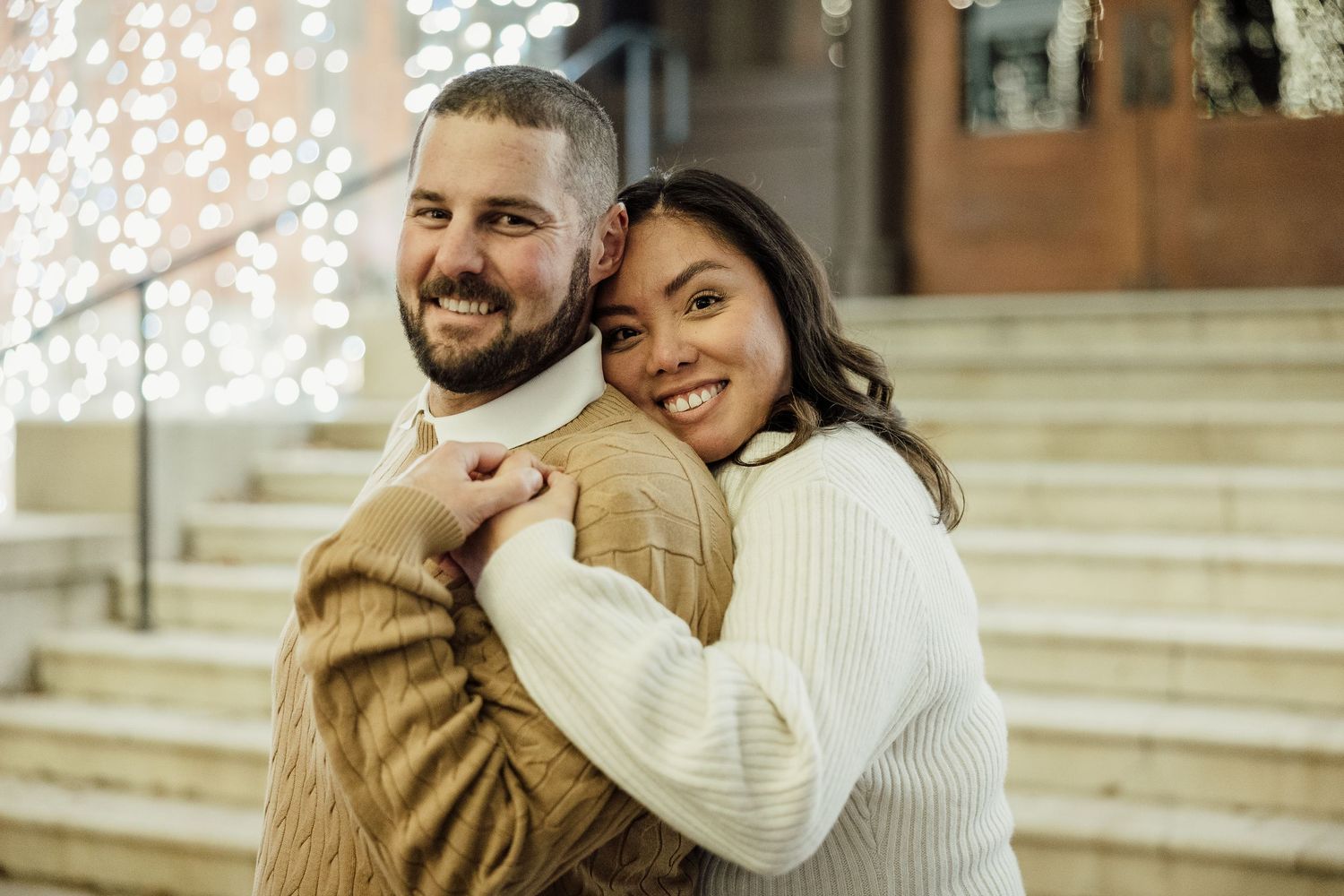 A couple in warm winter outfits embraces and smiles while standing on outdoor stone steps.