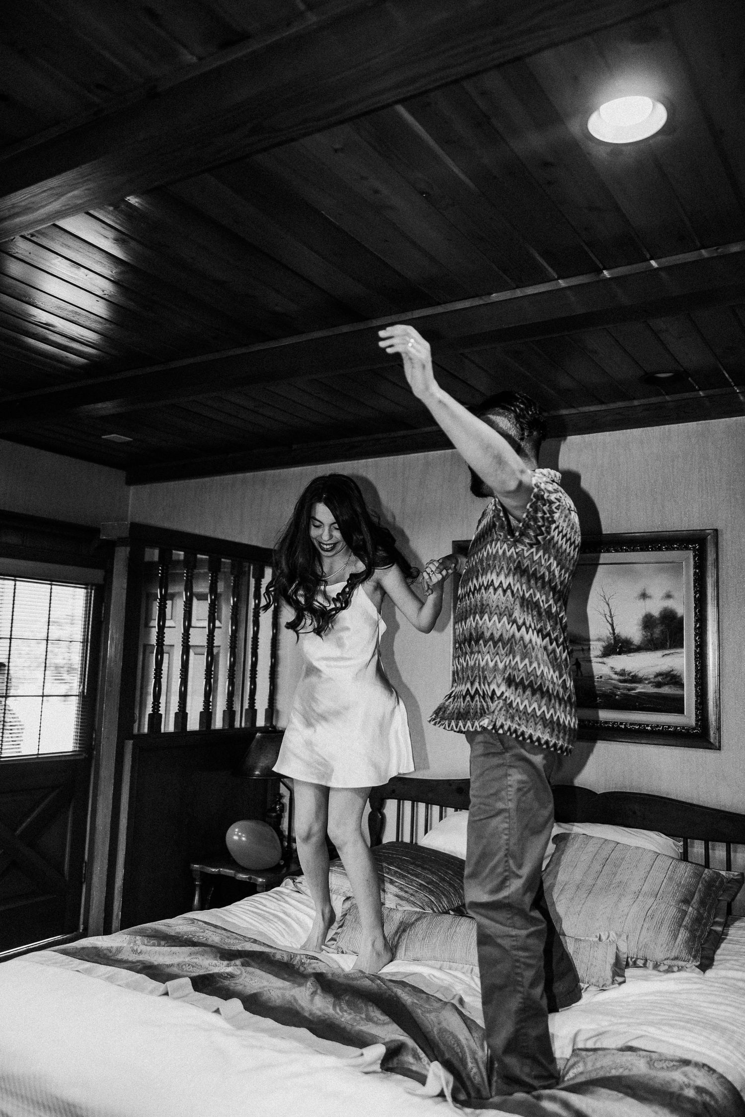 A couple joyfully jumps and dances together on a bed in a rustic bedroom with wood-paneled walls and ceiling.