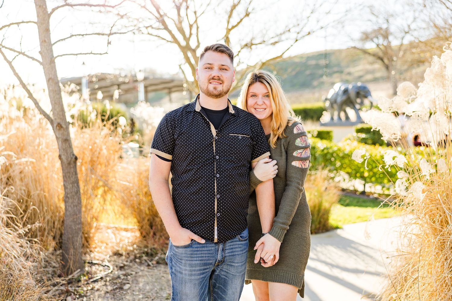 A couple poses together on a garden walkway with golden grass and trees in the background.