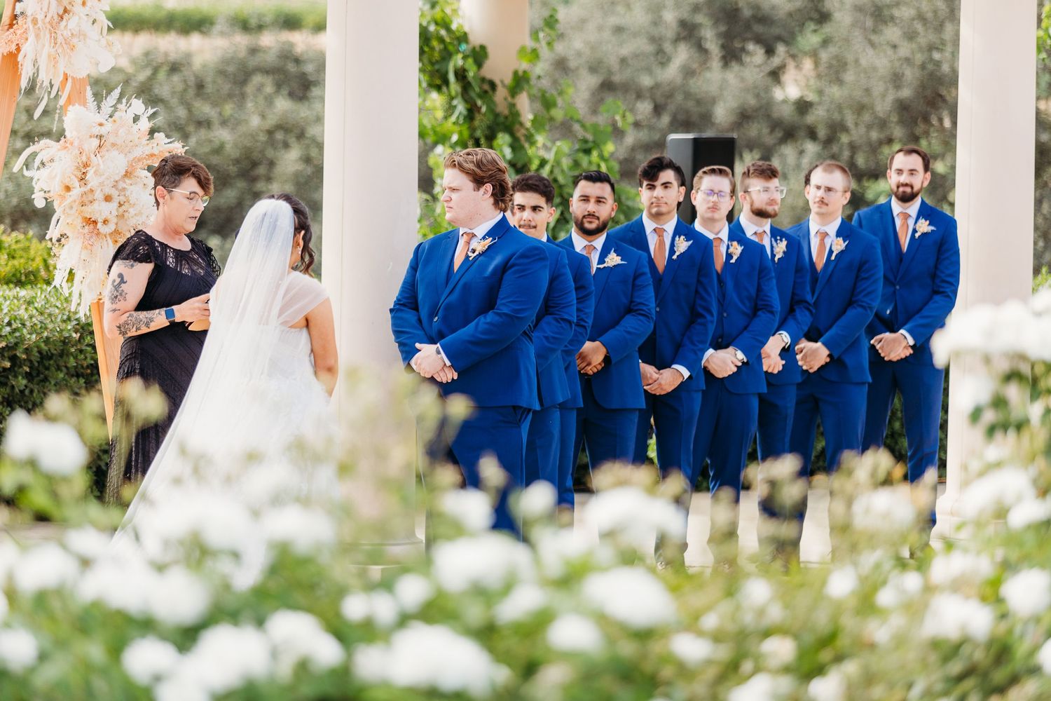 Wedding party in bright blue suits and white floral decorations gather during outdoor ceremony.