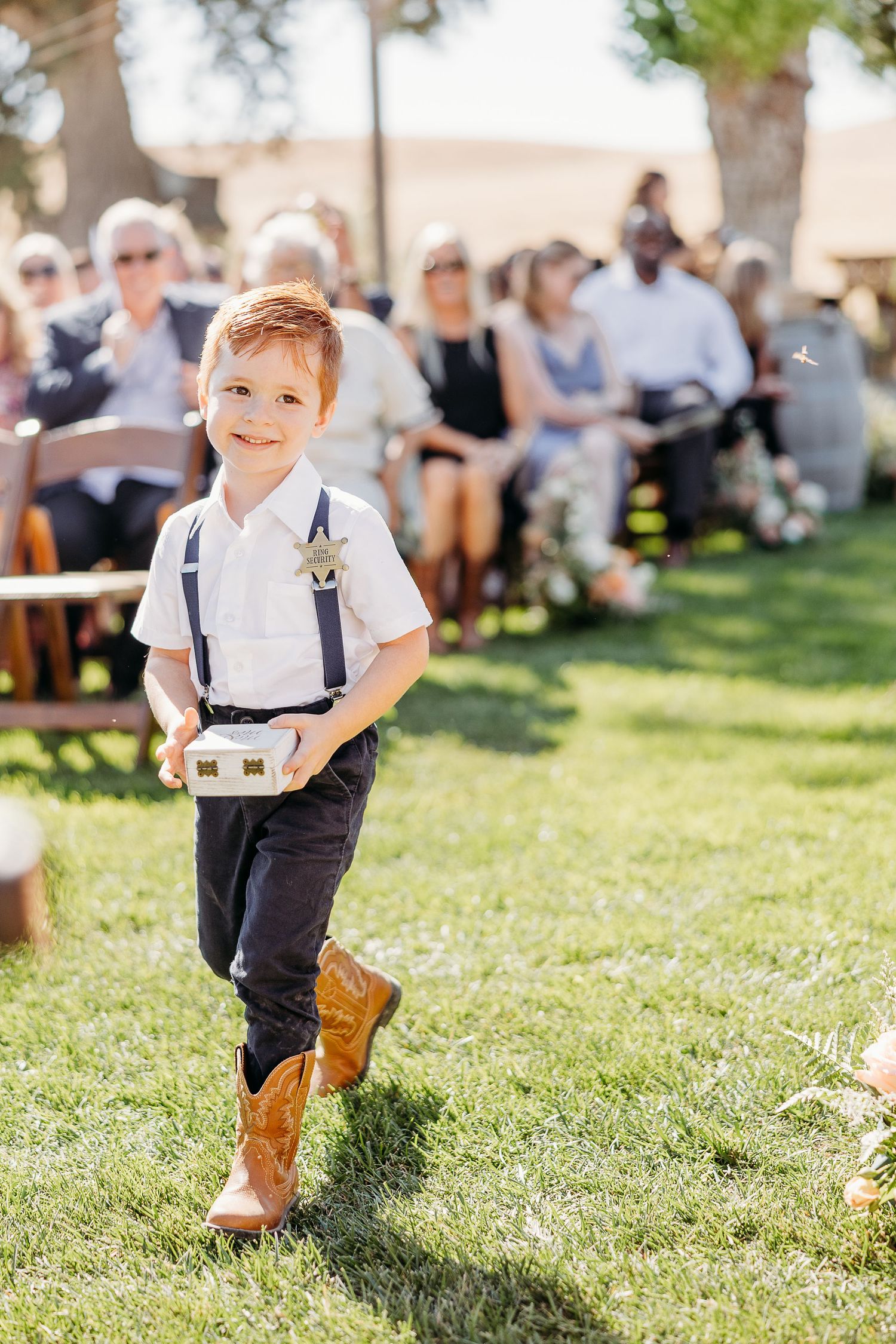 Young boy in suspenders and cowboy boots carries a ring box while walking on grass at an outdoor wedding ceremony.
