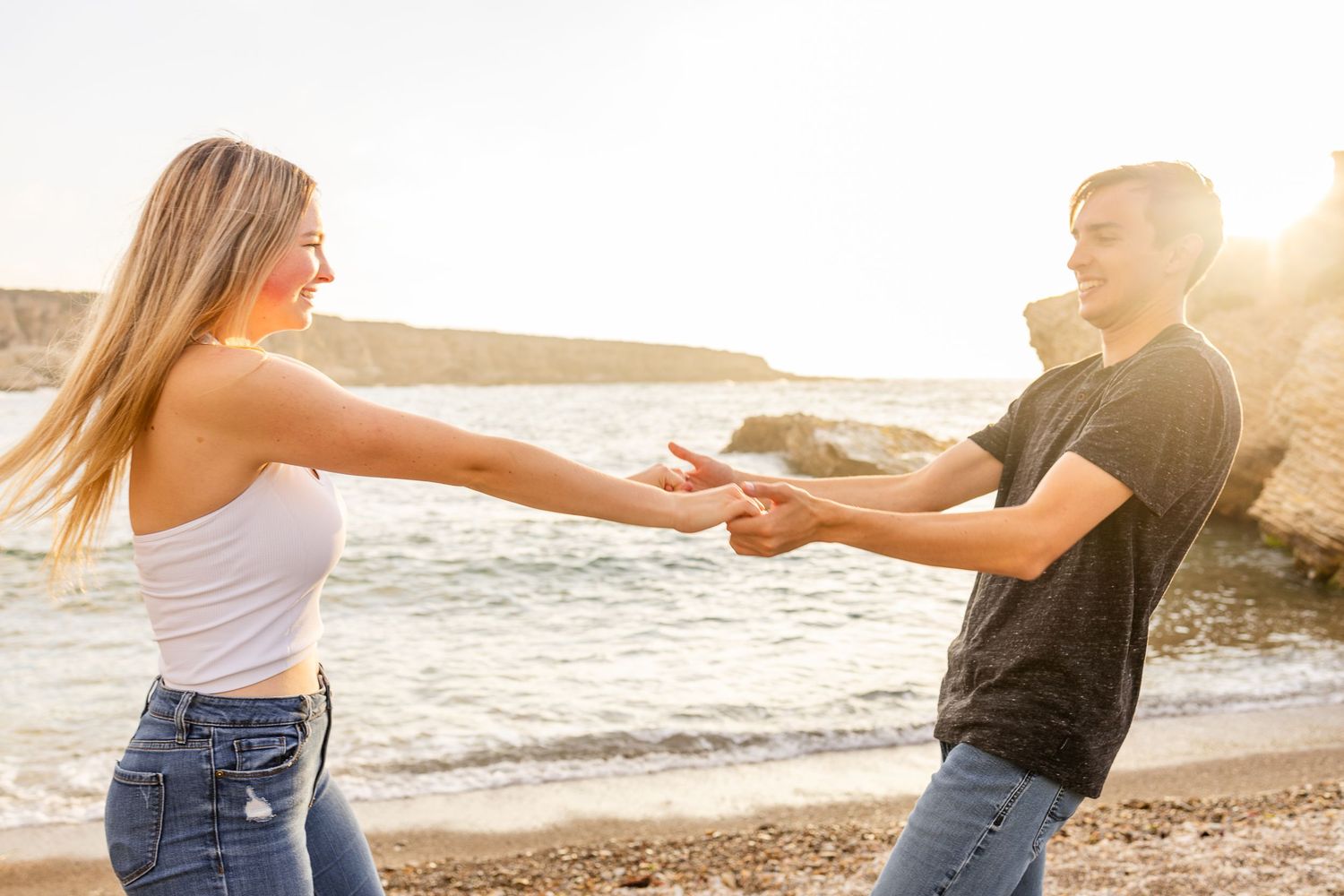 A couple holds hands while dancing on a beach with golden sunlight.