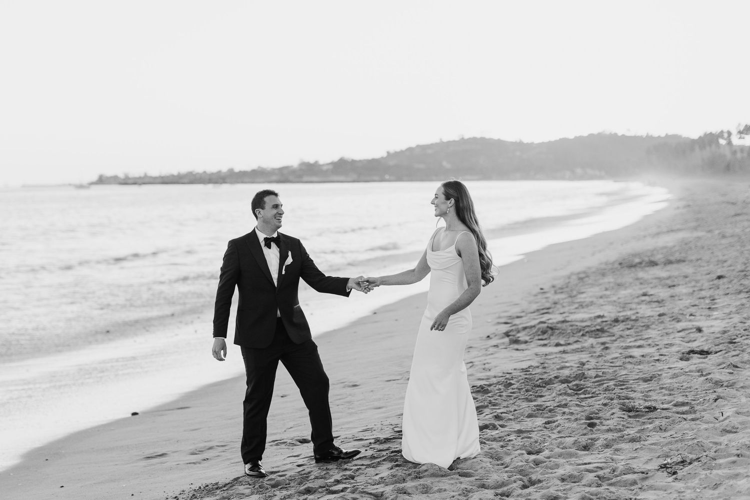 A black and white photo of a couple walking hand in hand along a scenic beach.
