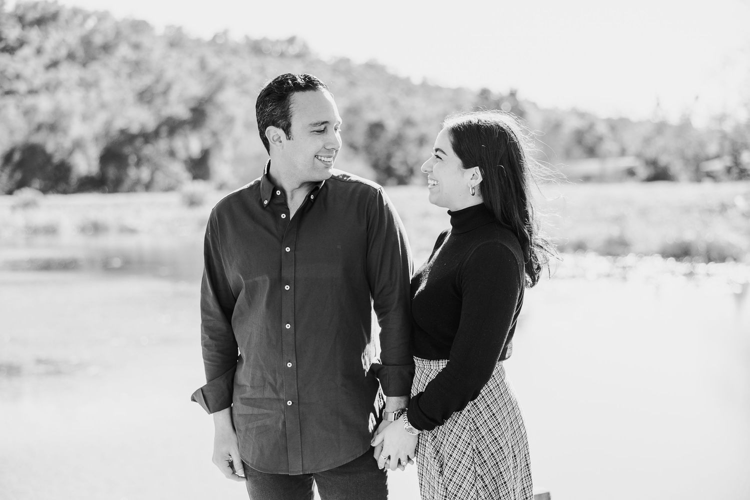 A couple shares laughter while posing together by a lake on a bright day.