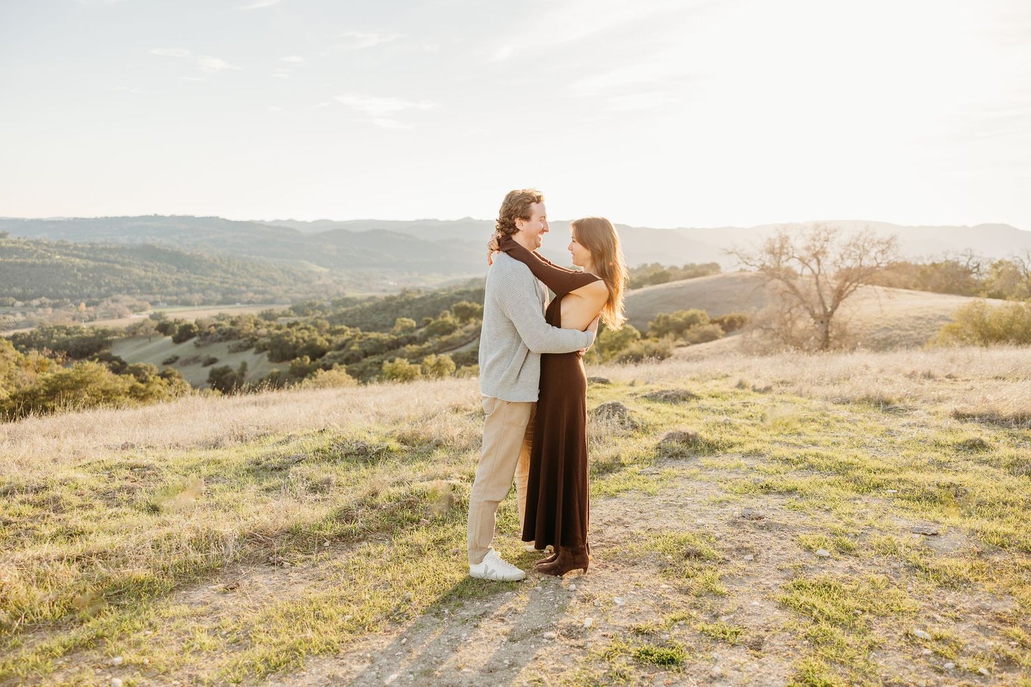 A romantic couple embraces on a hilltop during golden hour with rolling hills and mountains in the background.