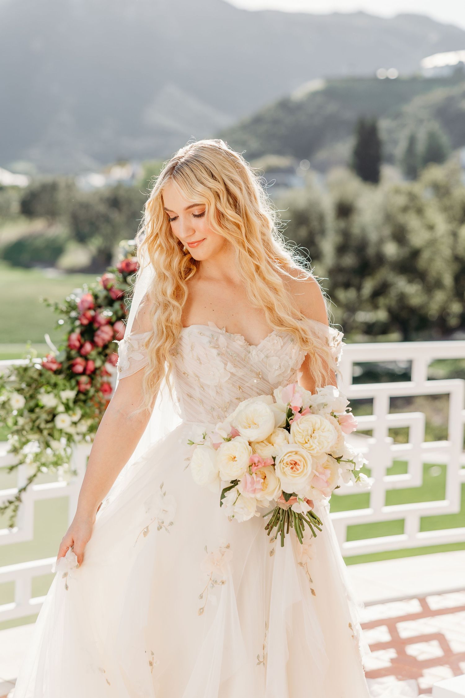 A bride in a lace gown poses with a pastel bouquet against a white fence and mountain backdrop.