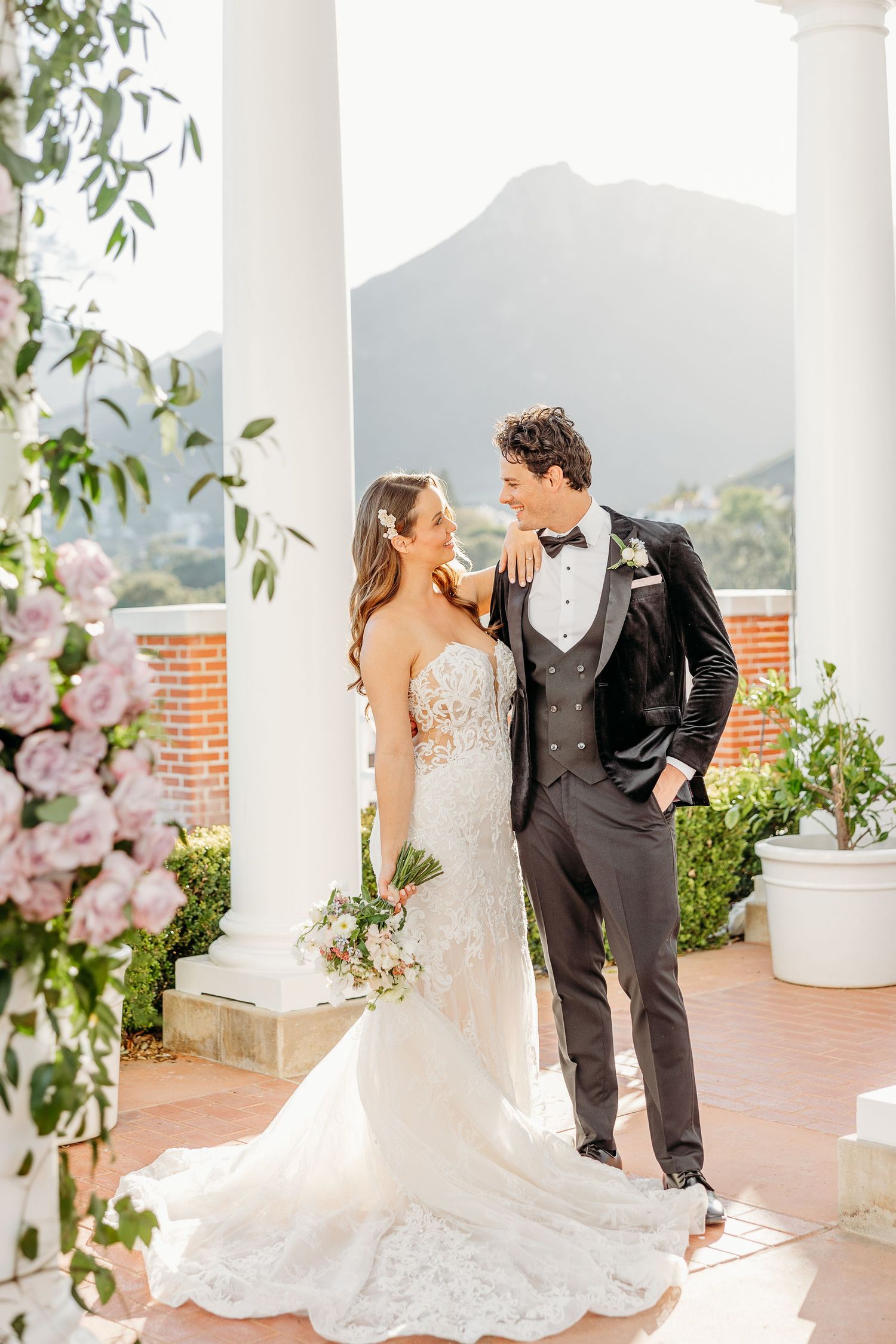 Elegant wedding couple shares romantic moments on a columned terrace with mountain views and floral decor.