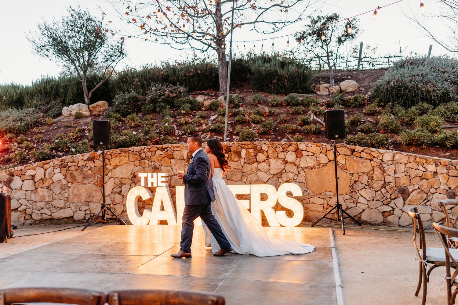 Illuminated marquee letters spell out text against a stone wall at an evening wedding venue.