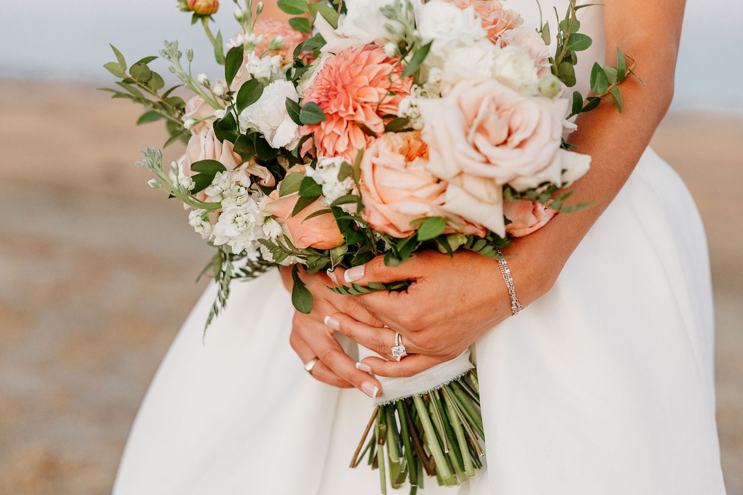 Elegant peach and white wedding bouquet with roses and greenery against a white dress.