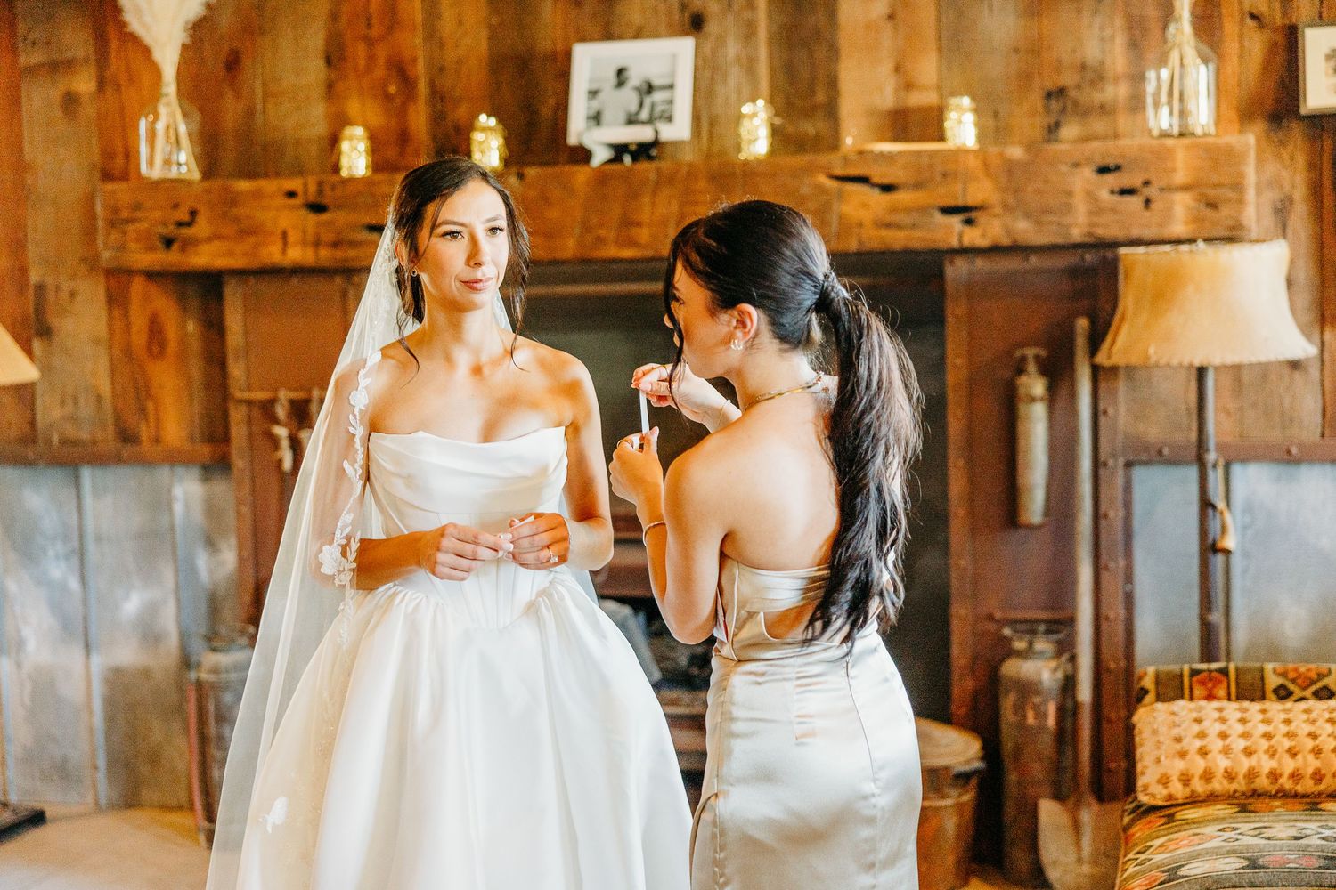 Two women in elegant dresses share a moment during a wedding celebration in a rustic wooden venue.