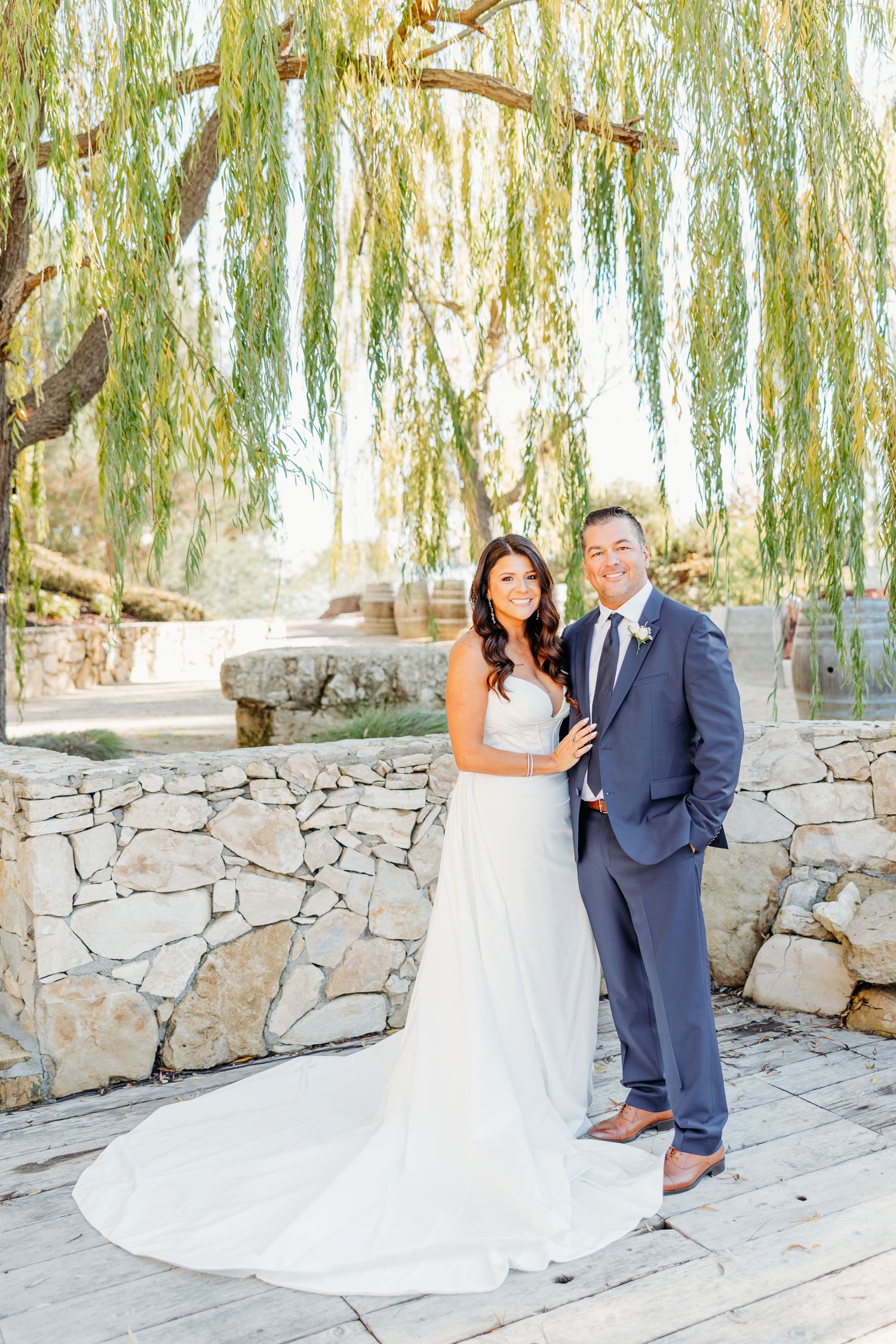 Wedding couple poses together against stone wall with romantic willow trees creating dreamy backdrop.