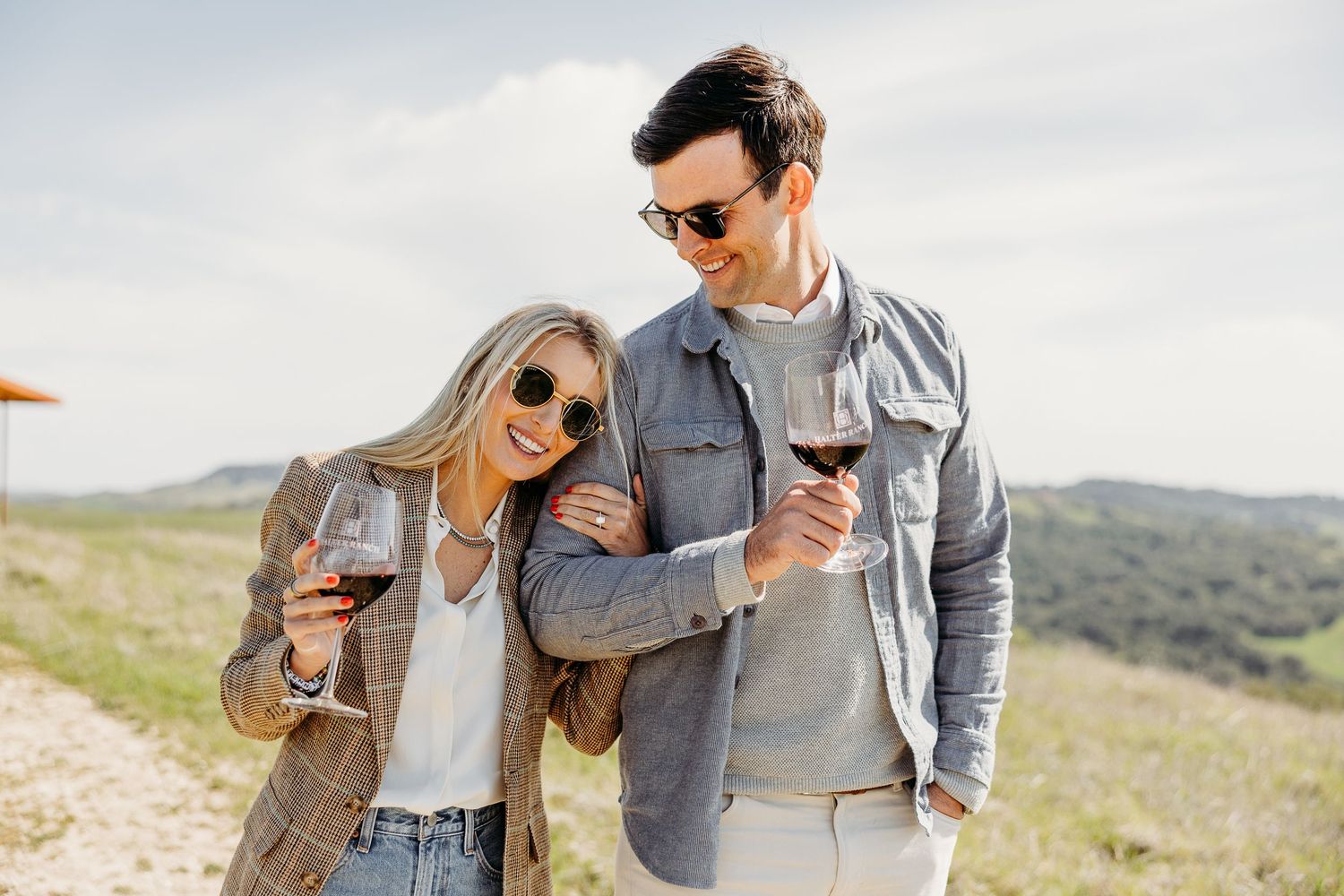 Couple in casual attire enjoying wine together outdoors in a scenic countryside setting.